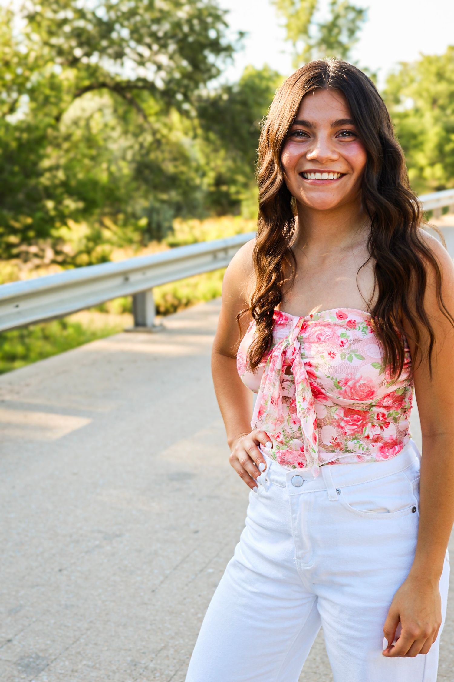 Smiling young woman in floral crop top and white jeans standing on a sunny tree-lined road.