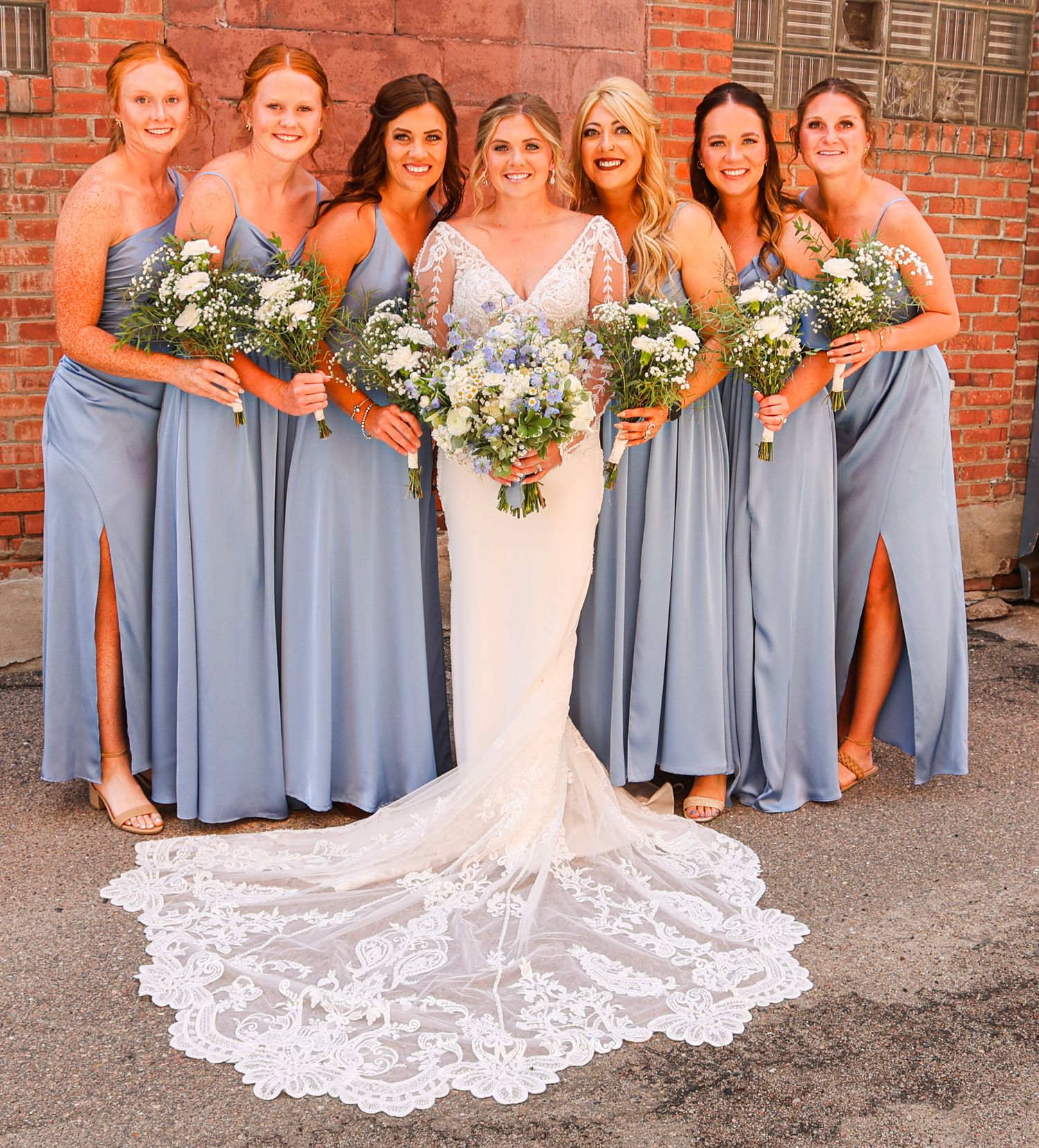 Bride in lace gown poses with four bridesmaids in dusty blue dresses holding wildflower bouquets.