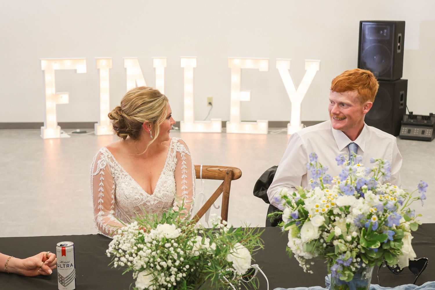 Bride and groom smile at sweetheart table with FINLEY marquee letters and blue floral centerpiece.