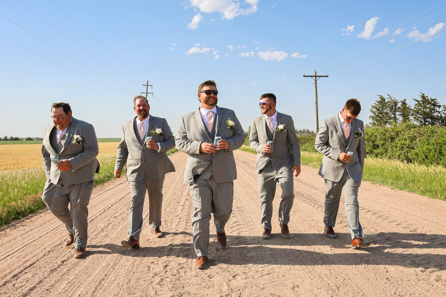 Five groomsmen in grey suits walking confidently down a sunny rural dirt road.