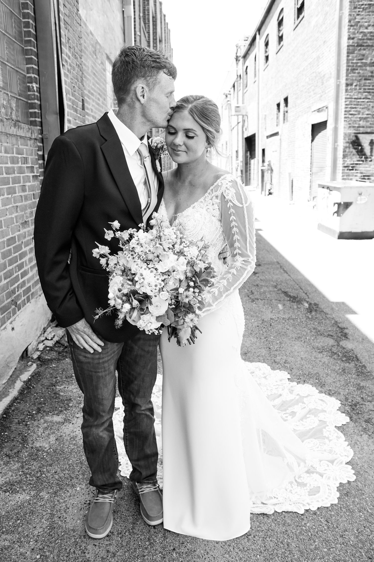 Black and white photo of a bride and groom sharing a tender moment in an urban alley, bride holding a floral bouquet.