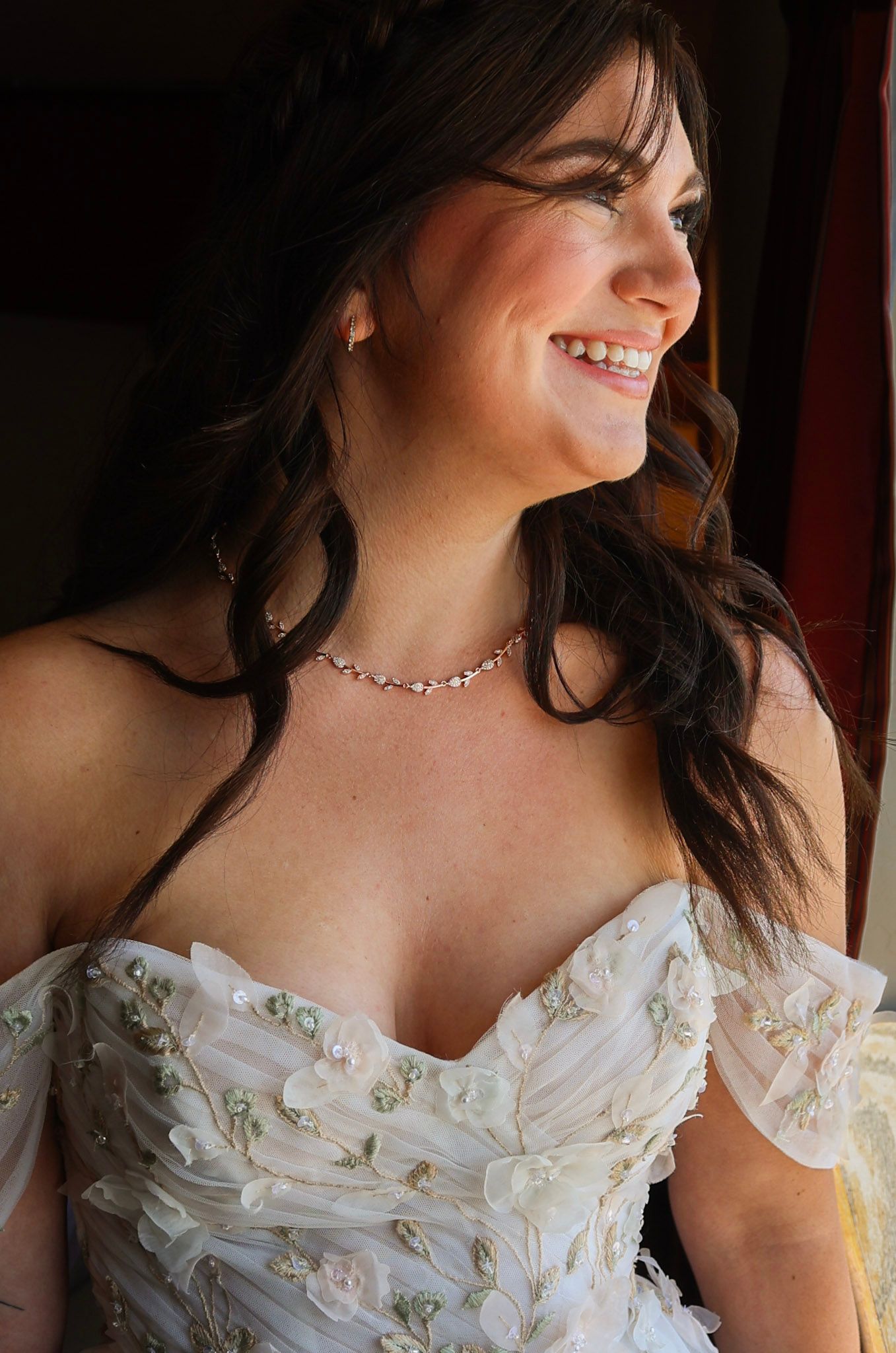 Smiling bride in floral off-shoulder white gown wearing a delicate necklace on her wedding day.