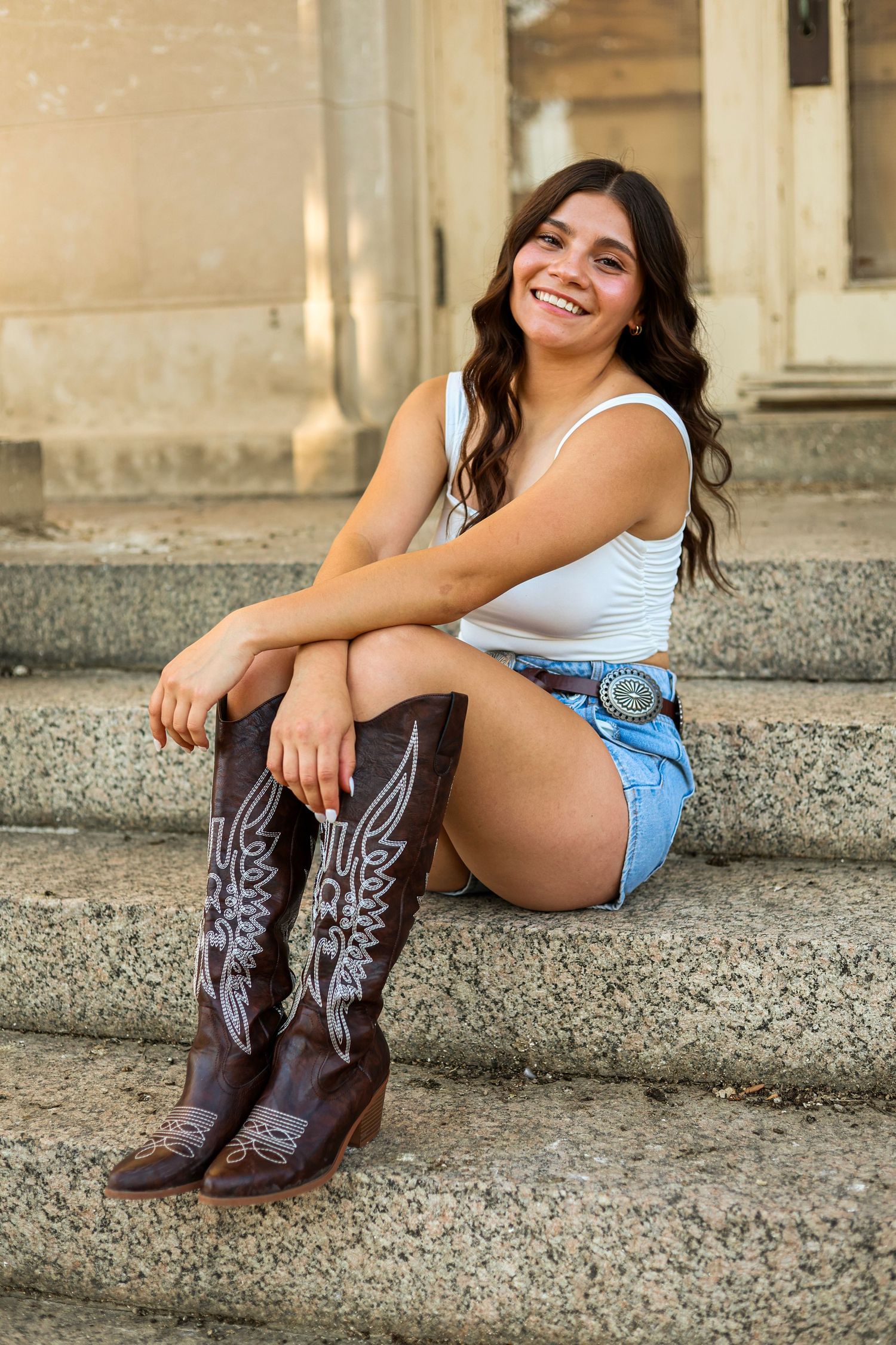 Smiling young woman in white tank top, denim shorts, and cowboy boots sitting on stone steps outdoors.