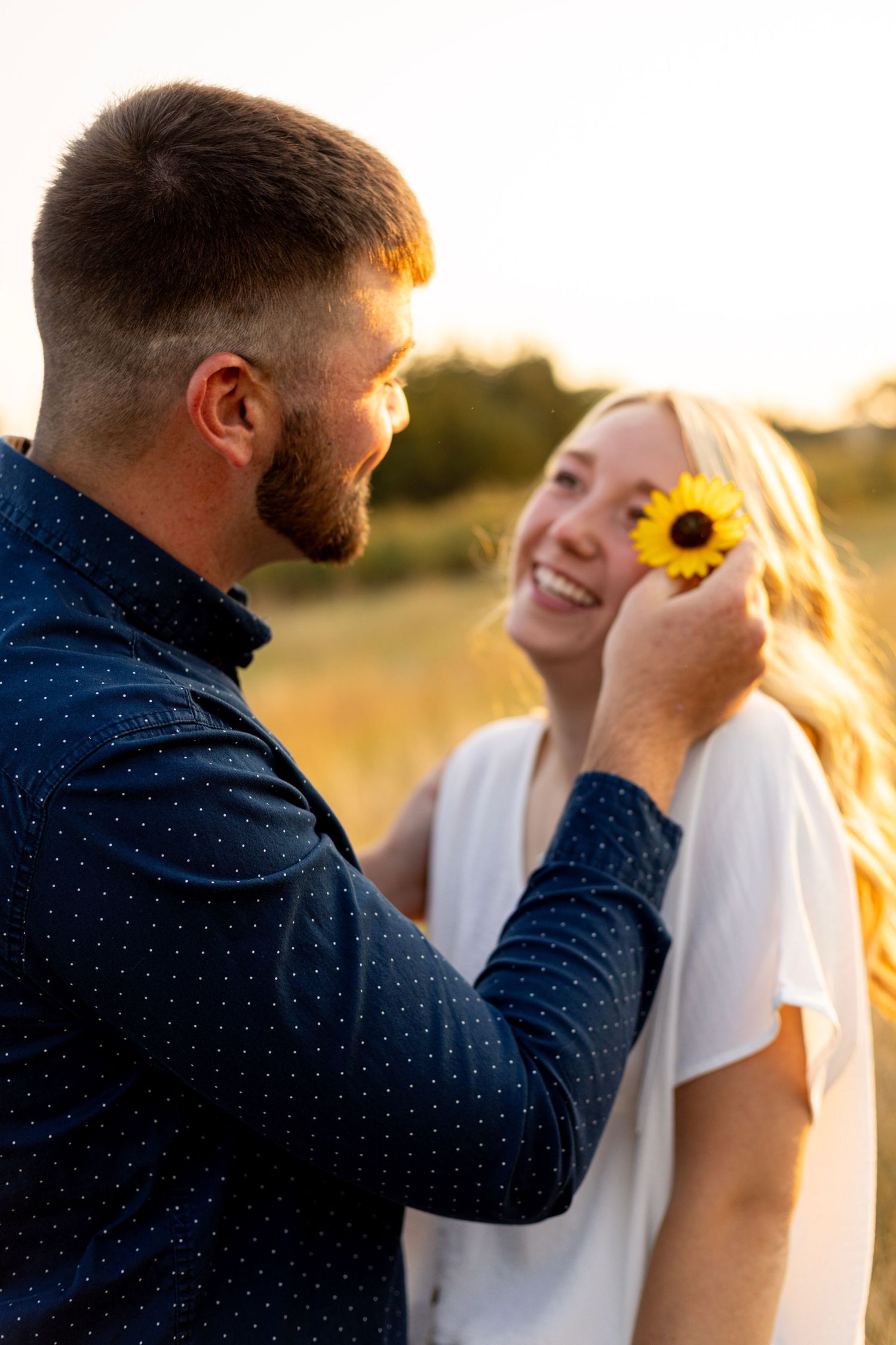Man holding a sunflower over smiling blonde woman's eye during a golden hour outdoor photo session.