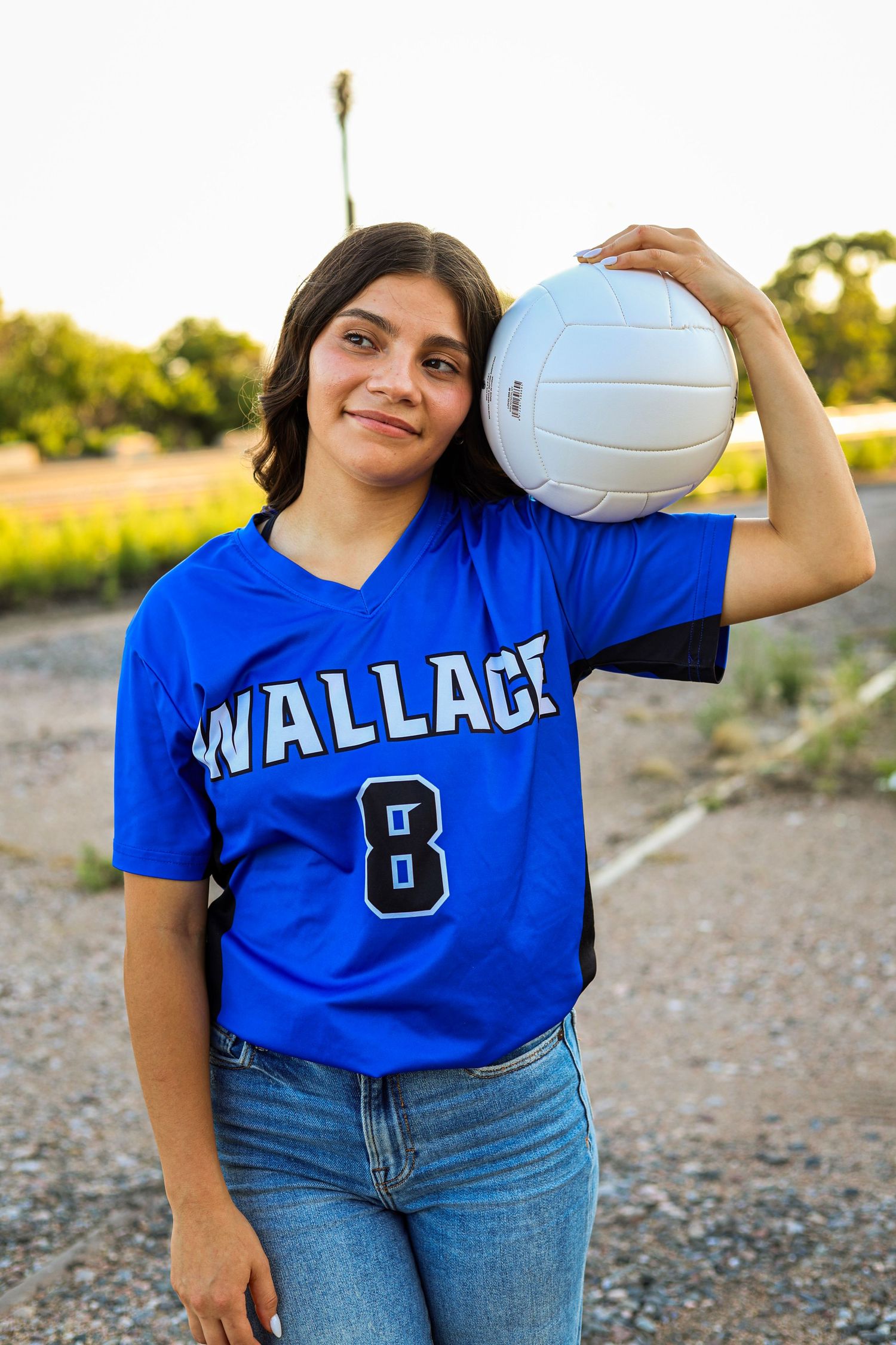 Young female volleyball player in blue Wallace number 8 jersey holding ball on rural dirt road.