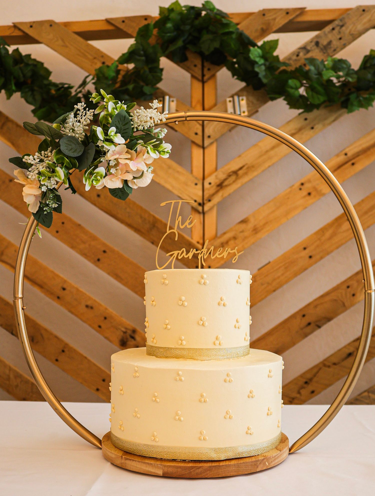 A two-tier white wedding cake displayed in a gold hoop stand with floral decor and The Gardners topper.