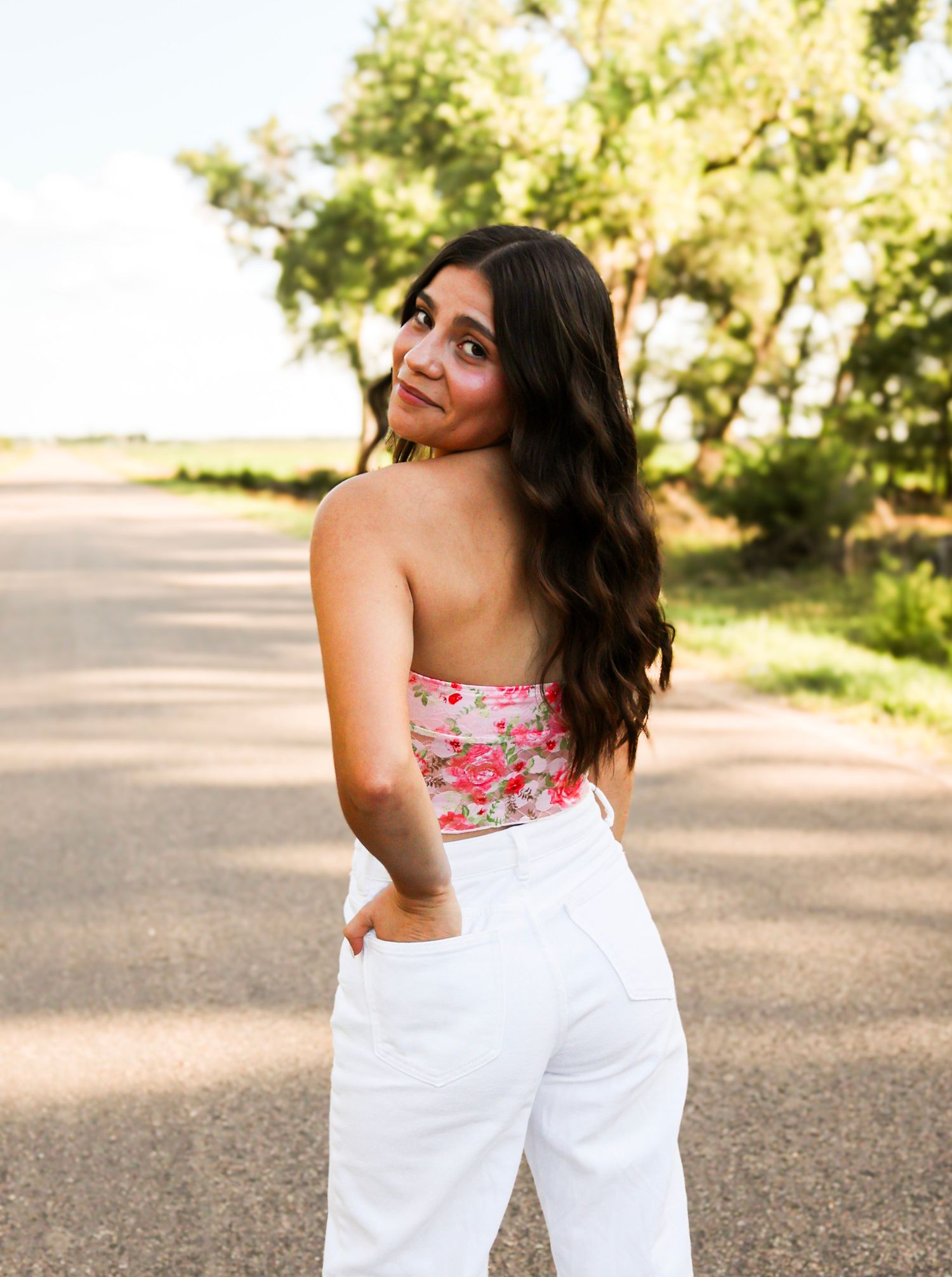 Young woman in floral tube top and white jeans looking back over her shoulder on a sunny rural road.