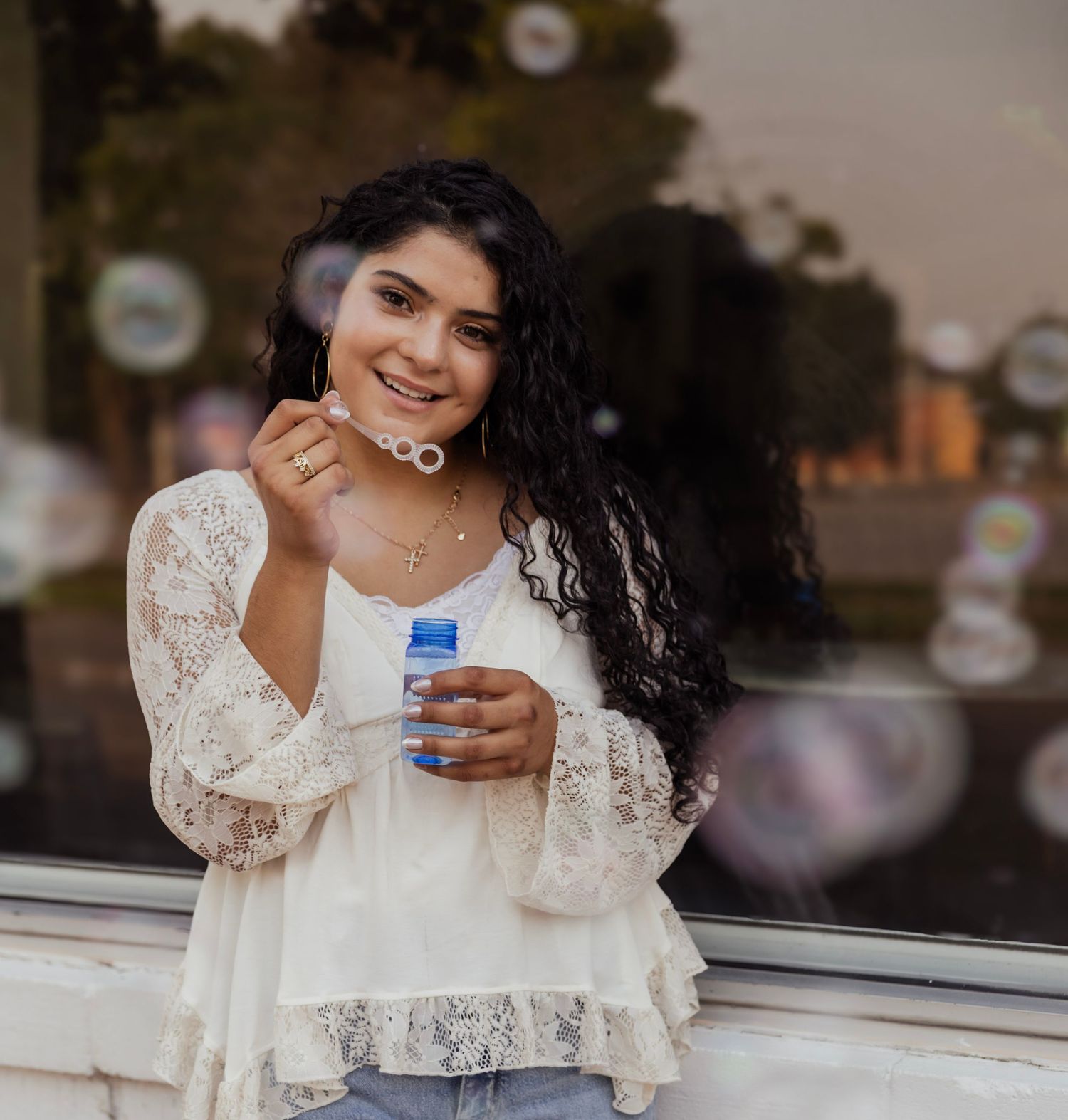 Young woman in white lace top blowing bubbles outdoors, smiling joyfully with curly dark hair.