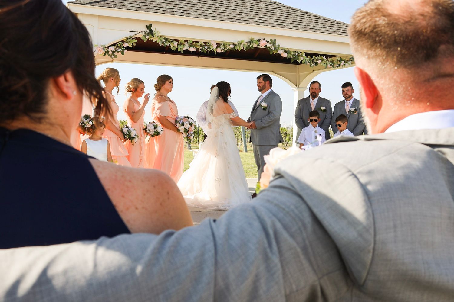 Wedding guests watch a ceremony at an outdoor gazebo decorated with flowers and a bridal party.