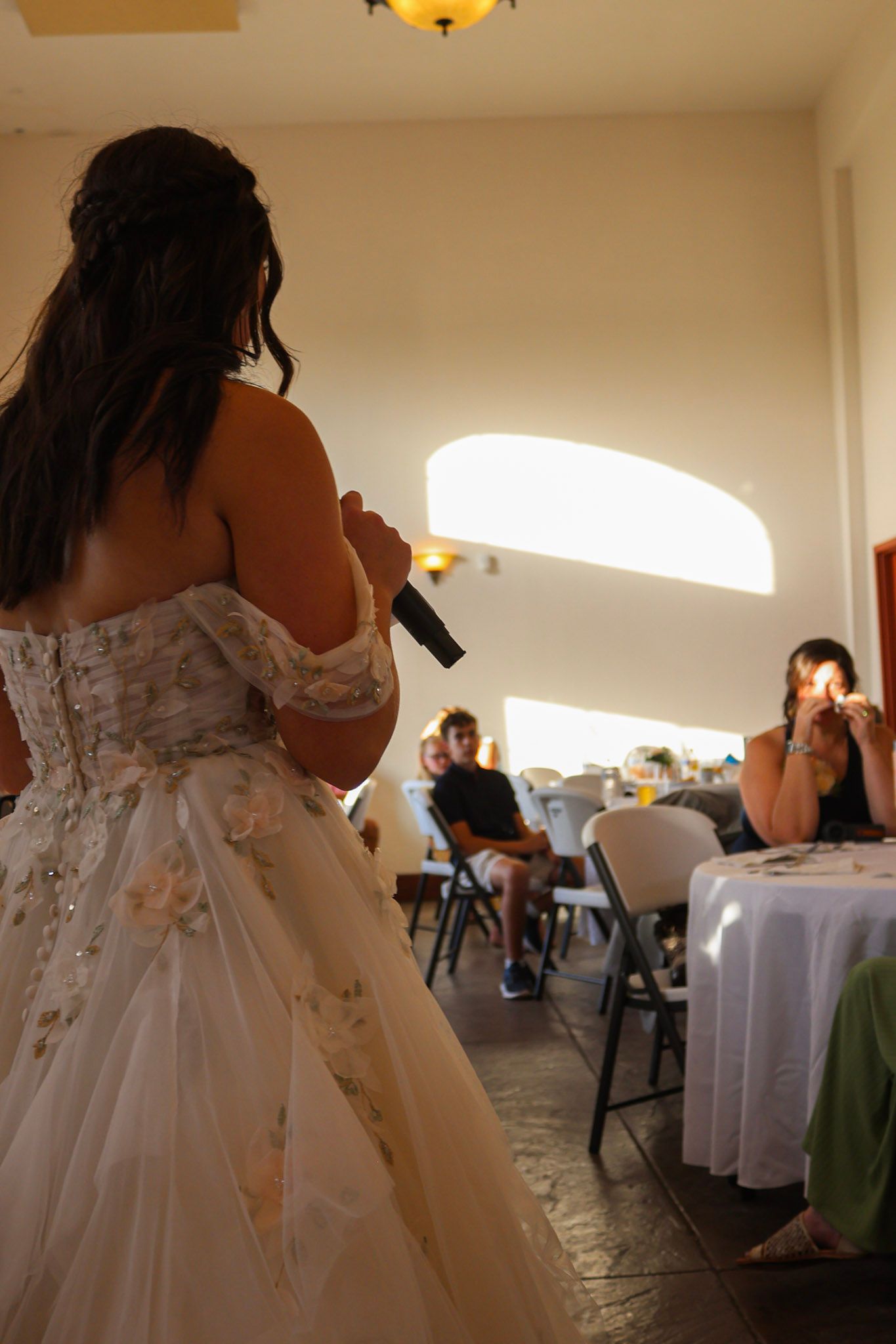 A bride in a floral off-shoulder gown speaks into a microphone during her wedding reception speech.