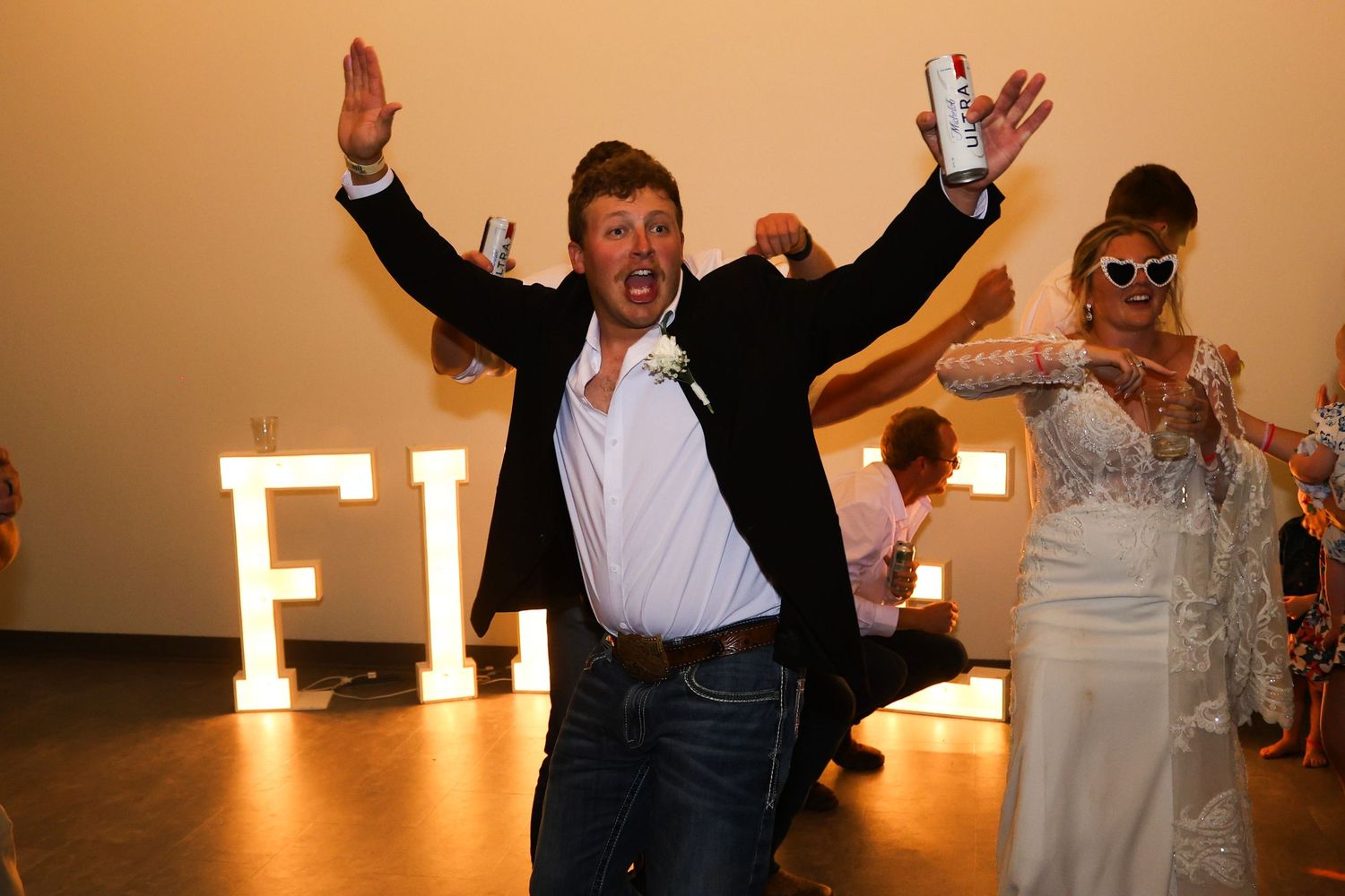 Excited wedding guest celebrates with arms raised beside bride in heart sunglasses at reception.
