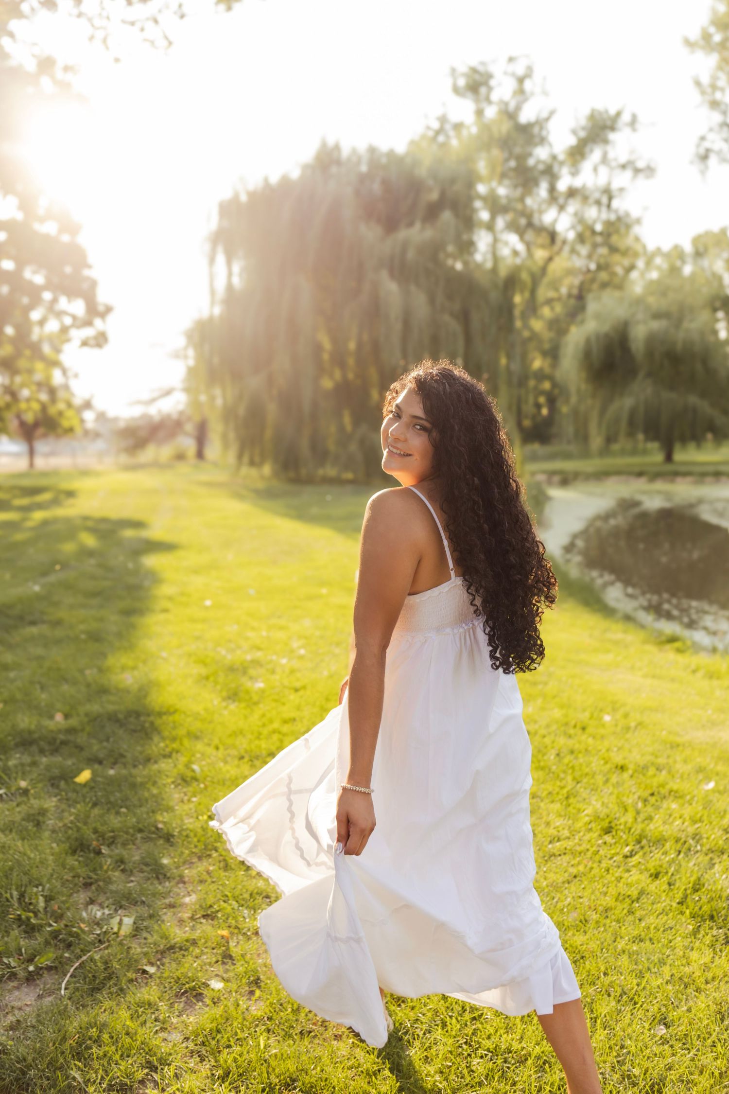 Young woman in white sundress twirling in sunny park by pond with willow trees, long curly hair flowing.
