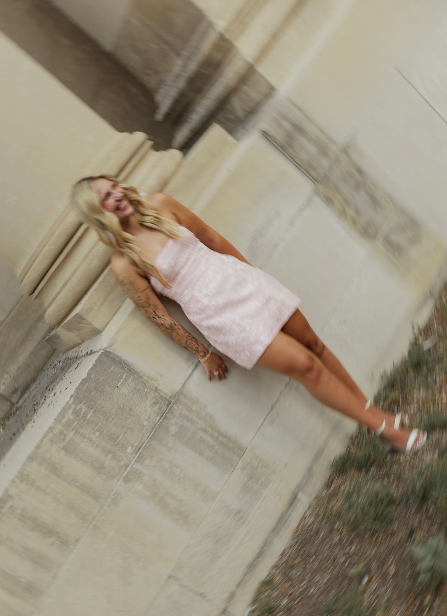 Young blonde woman in a pink mini dress and white heels posing outdoors near stone columns.