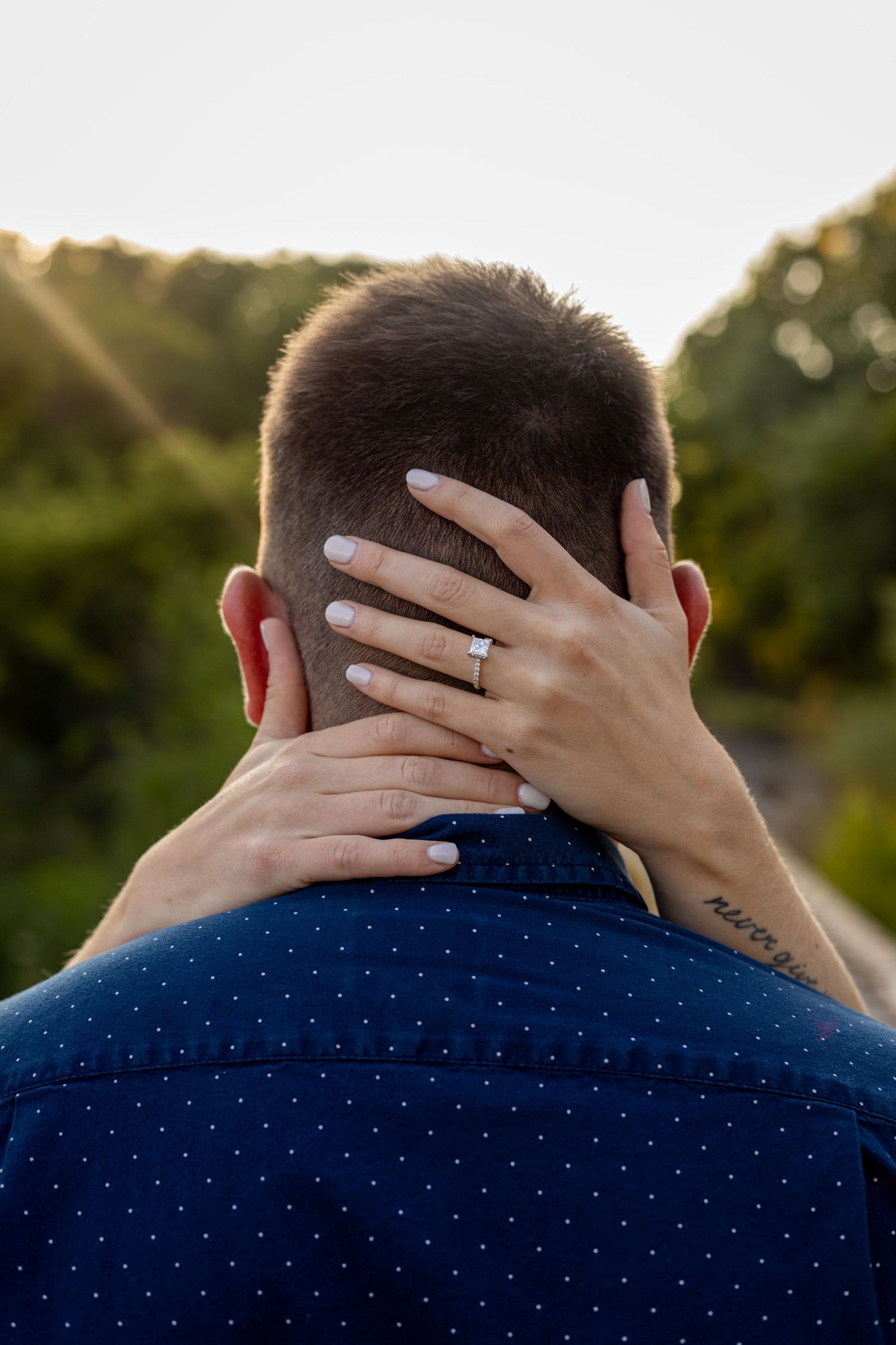 Woman with engagement ring and wrist tattoo tenderly cradles mans head from behind at golden hour.