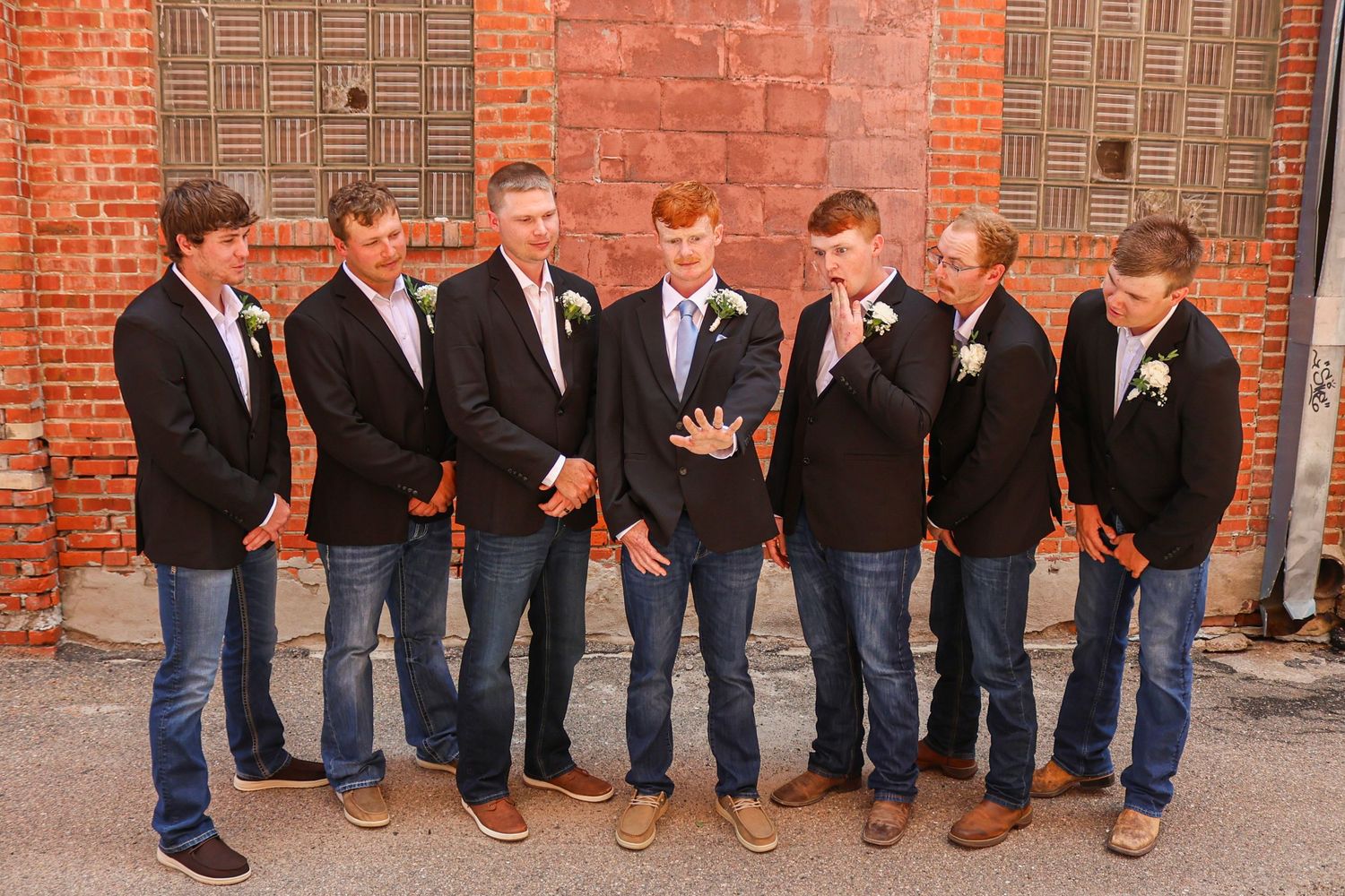 Seven groomsmen in navy blazers and jeans pose together outdoors against a brick wall at a wedding.