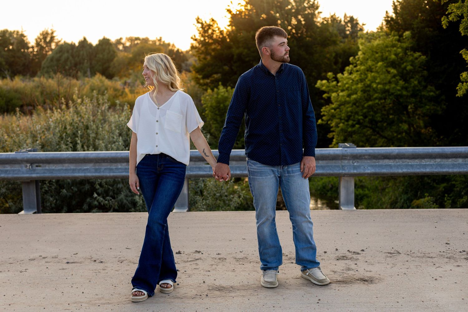 An engaged couple walks hand in hand on a sandy path at sunset near a bridge.