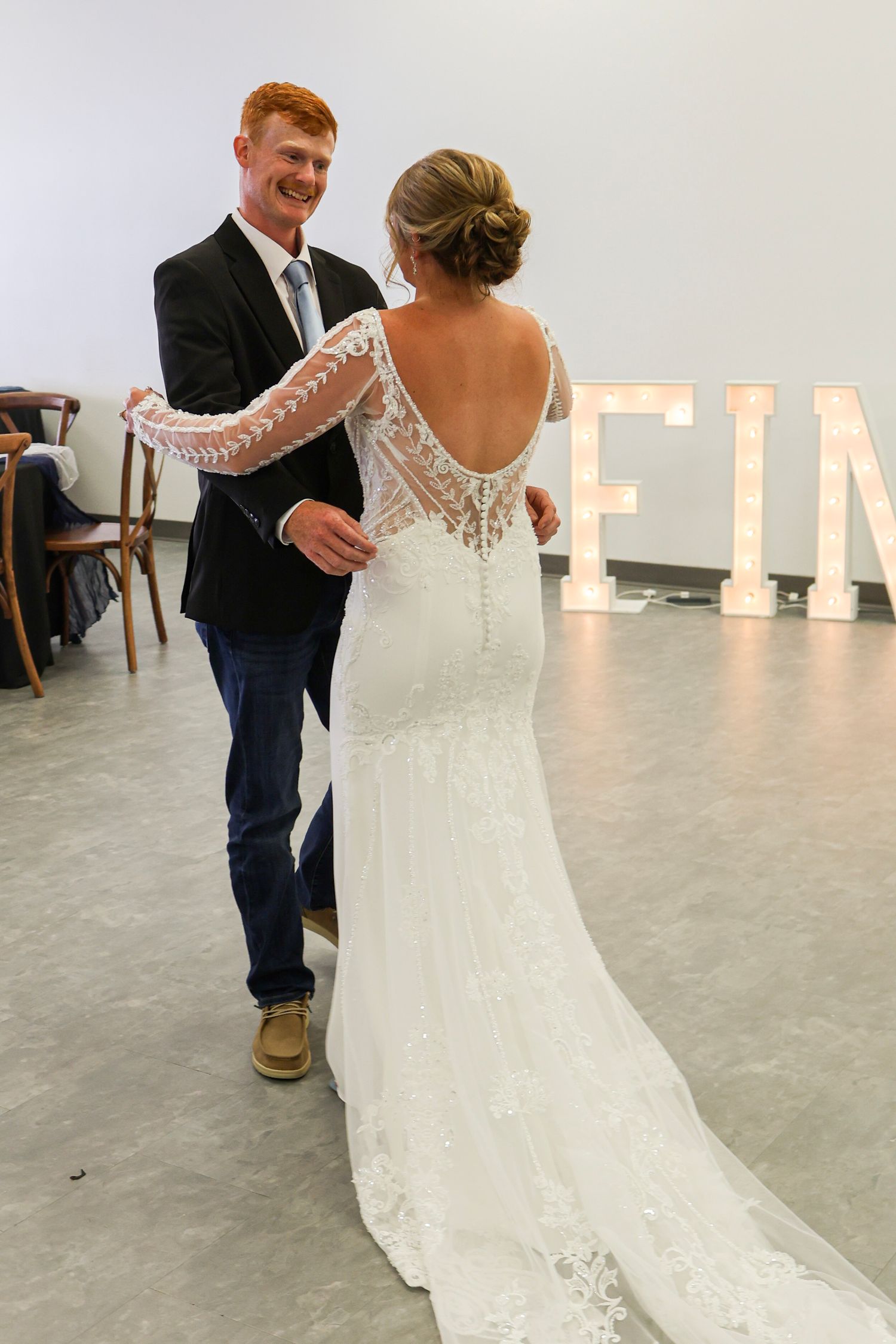 Redhead groom in black suit smiles at bride in backless lace wedding gown near FINLEY marquee letters.