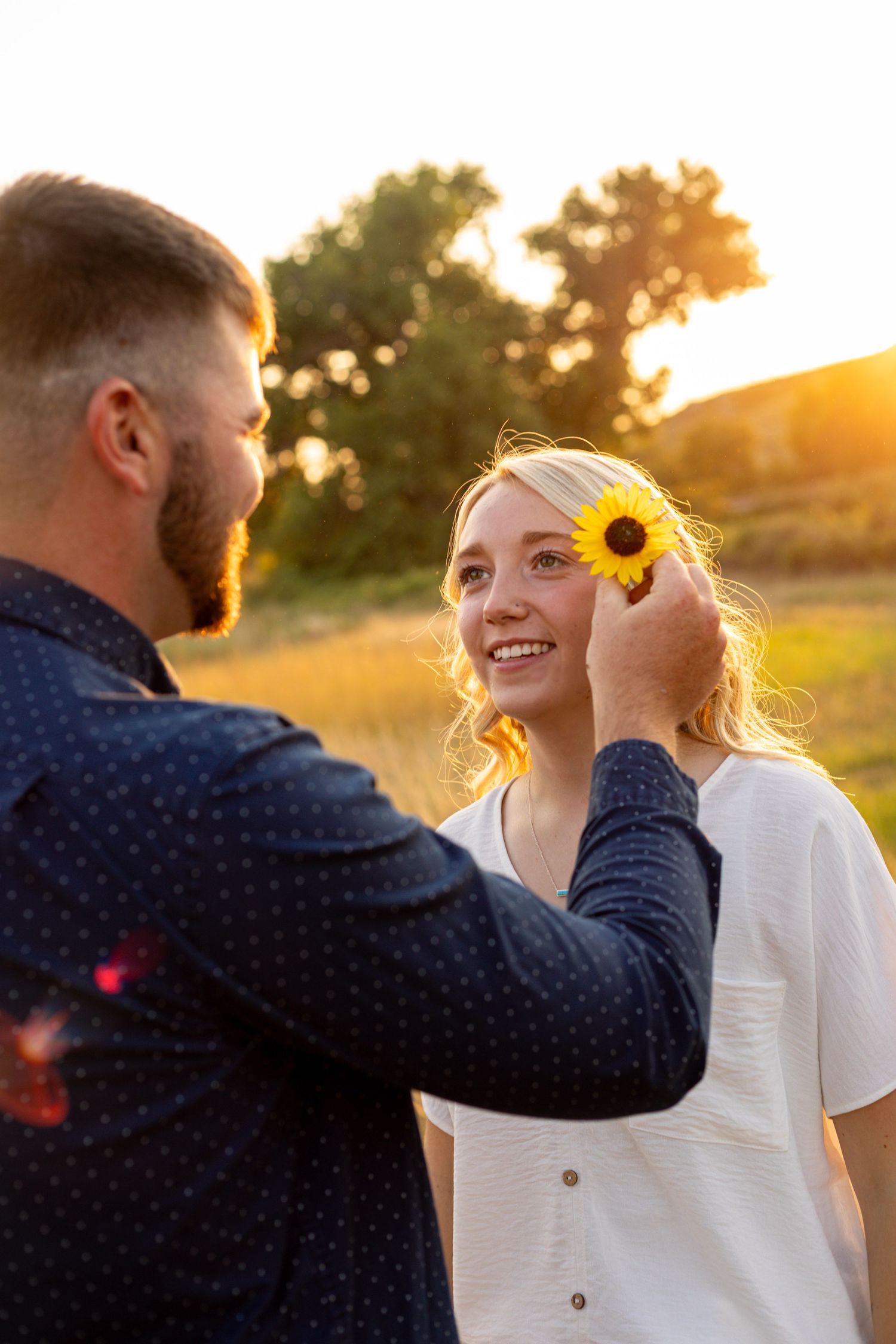 A man places a sunflower in his smiling girlfriends hair during a warm golden sunset outdoors.