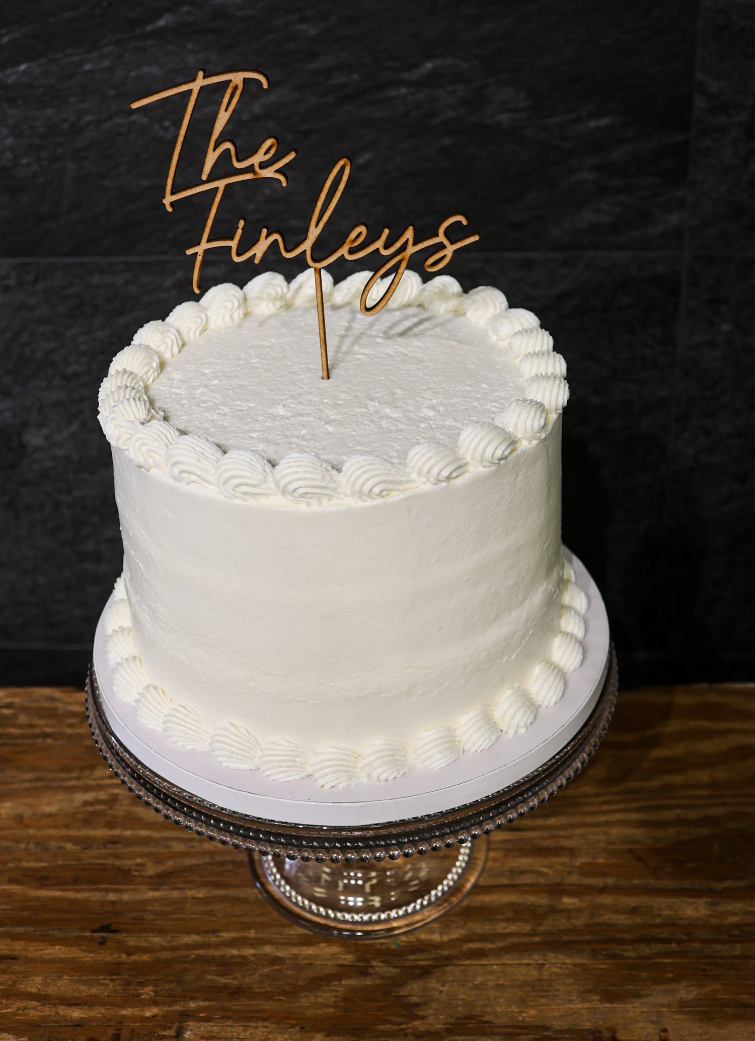 Elegant white wedding cake with rosette border and The Finleys wooden cake topper on glass stand.