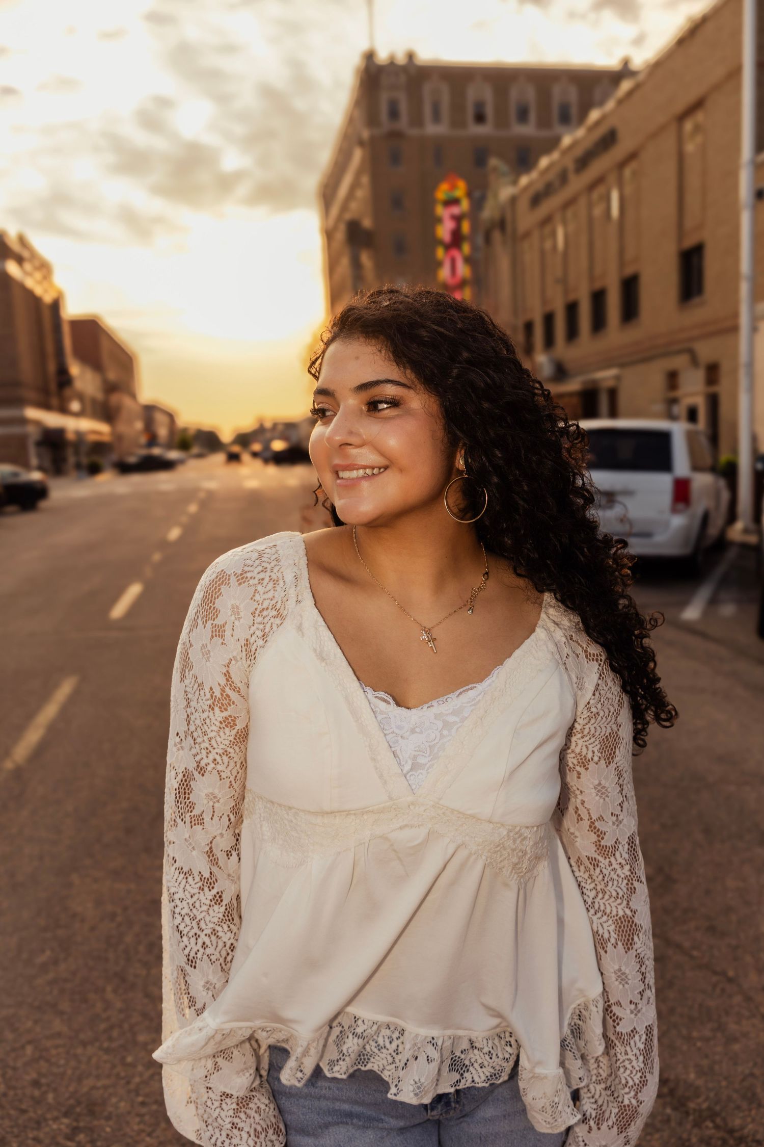Young woman with curly hair smiles in white lace top on a city street at golden hour sunset.