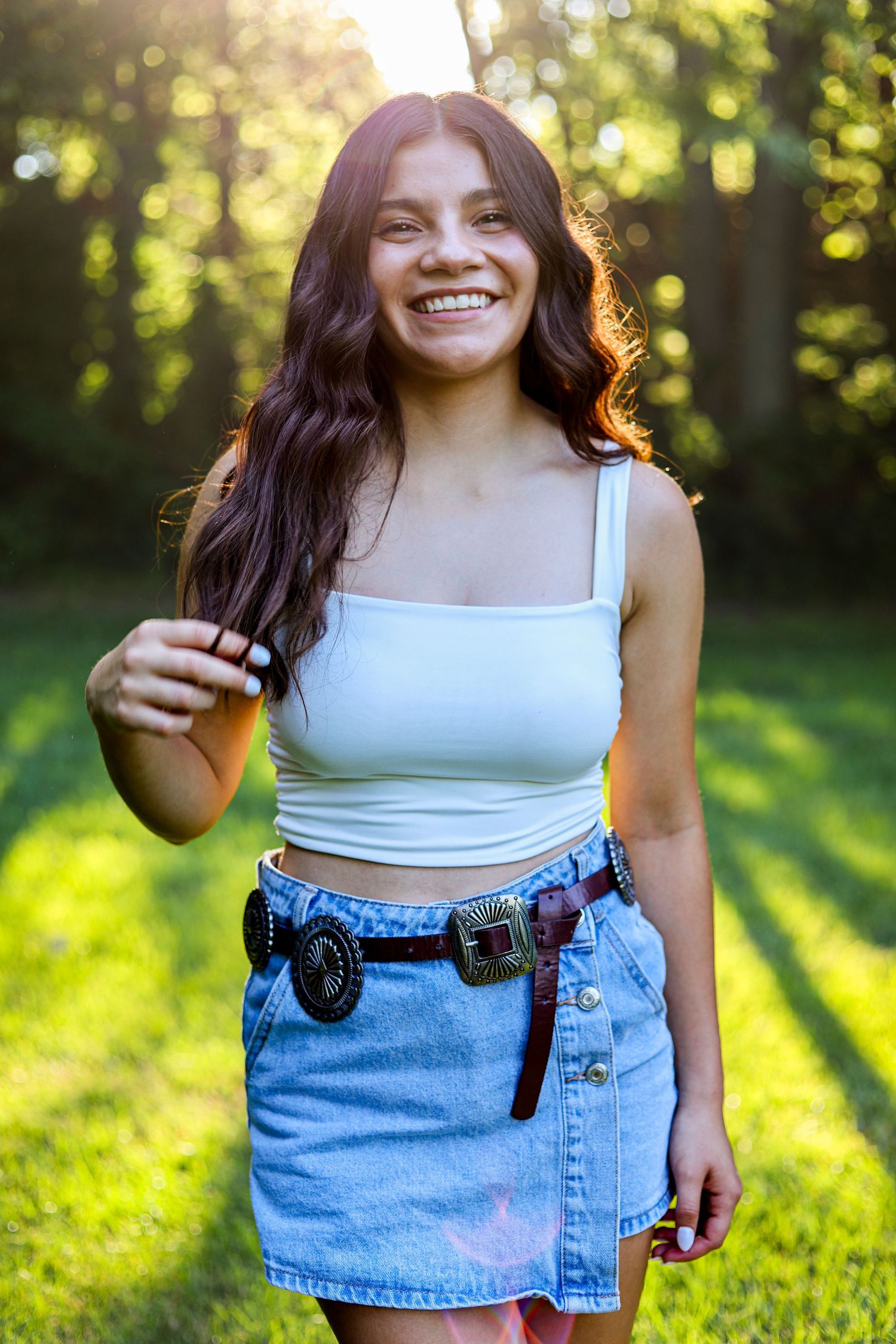 Happy young woman in white crop top and denim skirt with concho belt posing in sunlit green park.