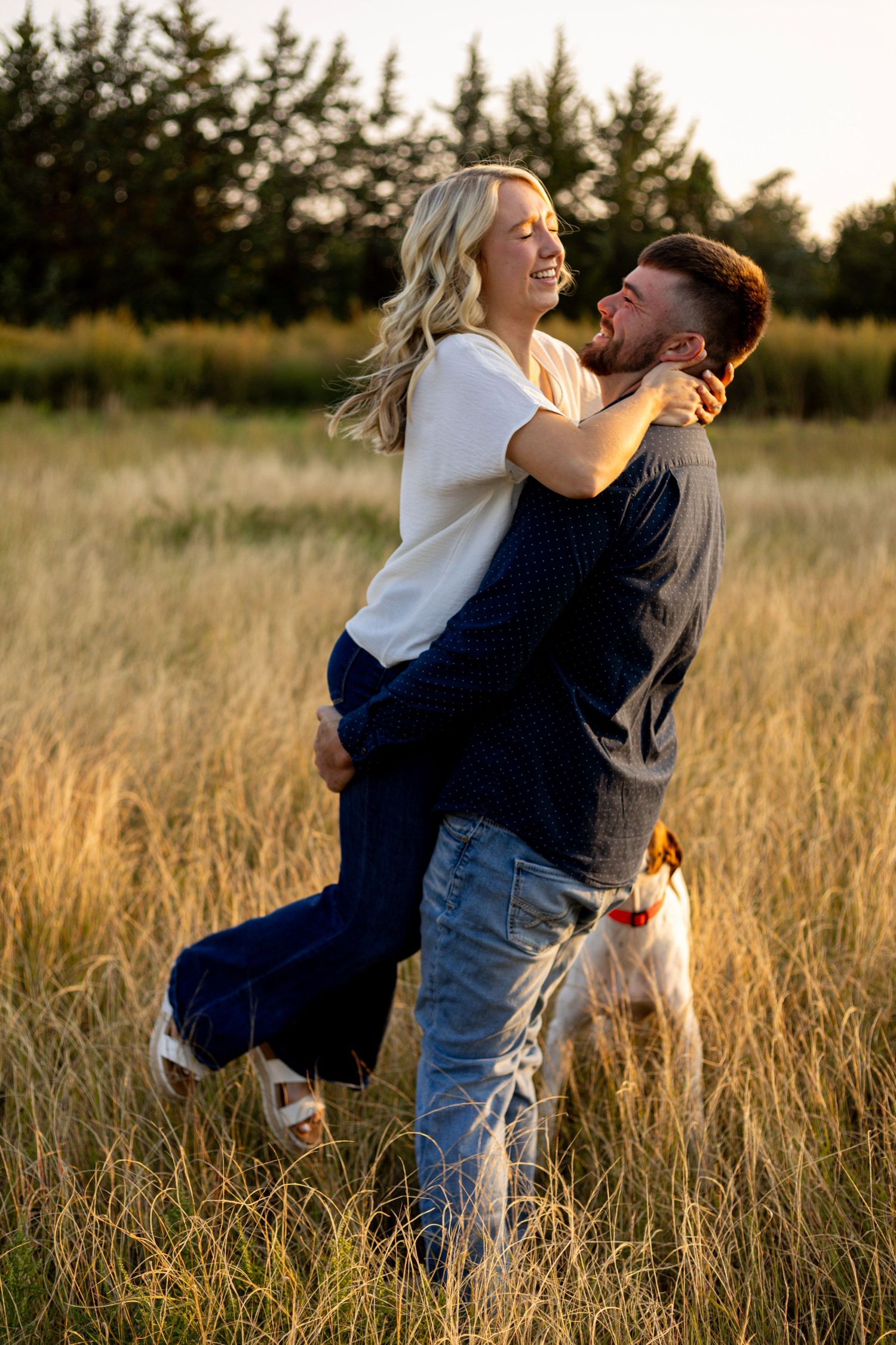 A joyful man lifts his laughing fiancee in a golden wheat field at sunset with a dog nearby.