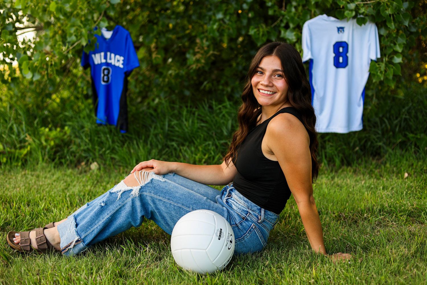 Teen girl in black top sitting on grass with volleyball and two jerseys hanging in trees behind her.