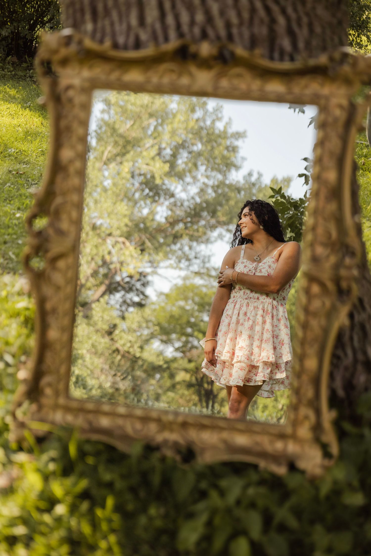 A young woman in a floral dress reflected in an ornate gold mirror outdoors surrounded by lush green trees.