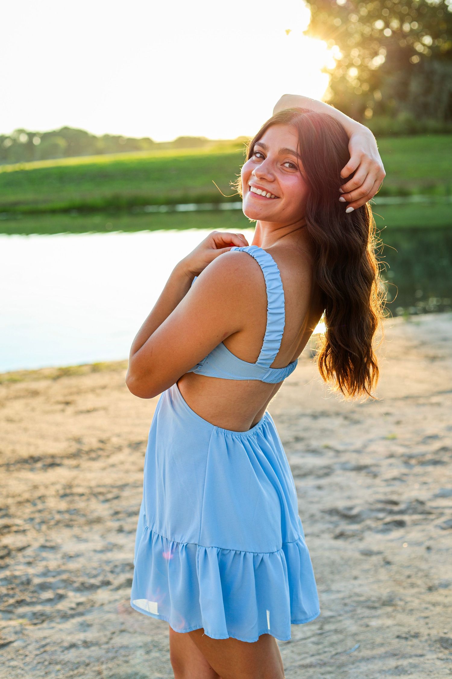 Young woman in backless light blue dress smiling over shoulder at sandy lakeside during golden sunset.