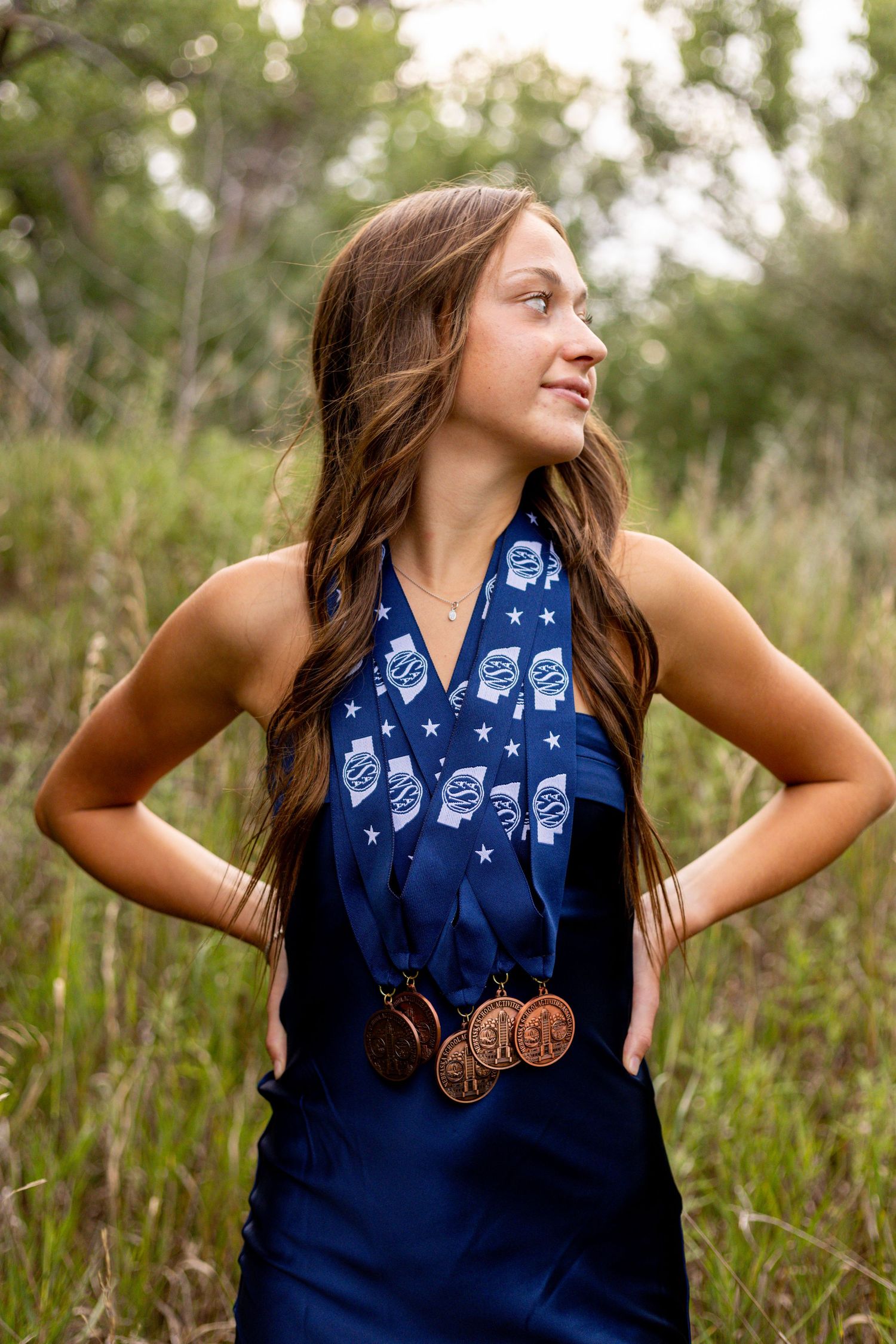 Young woman in navy halter dress proudly wearing bronze medals and patriotic ribbon in natural setting.