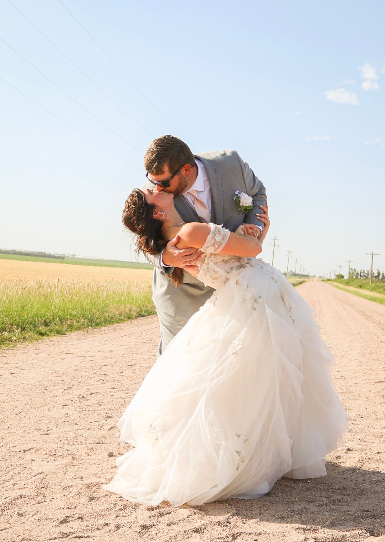Bride and groom sharing romantic dip kiss on a sunny rural dirt road.