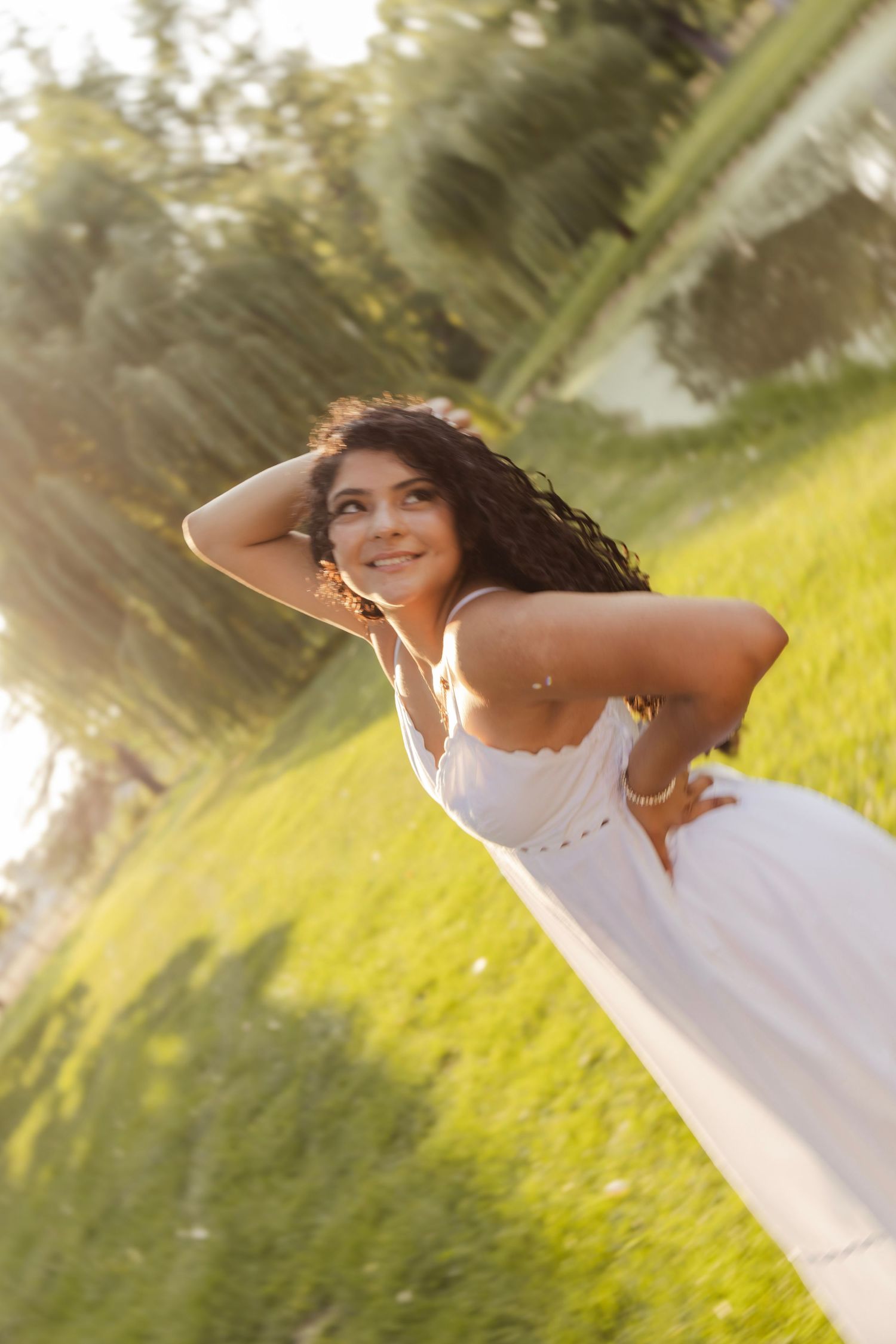 Young woman in white dress smiling outdoors in golden sunlight, with flowing dark hair and green grass background.