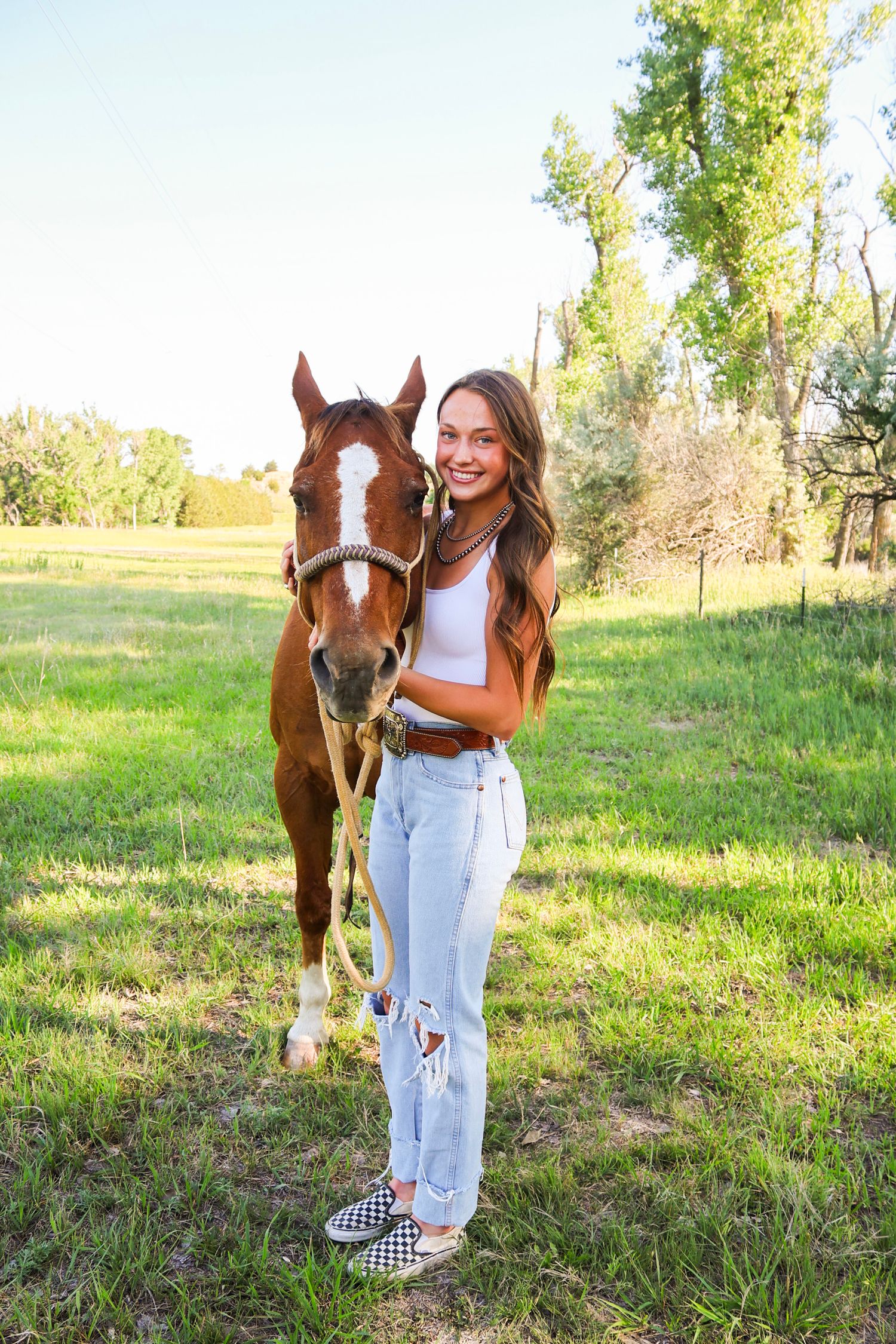 Smiling teenage girl in ripped jeans posing with a chestnut horse in a sunny green pasture.