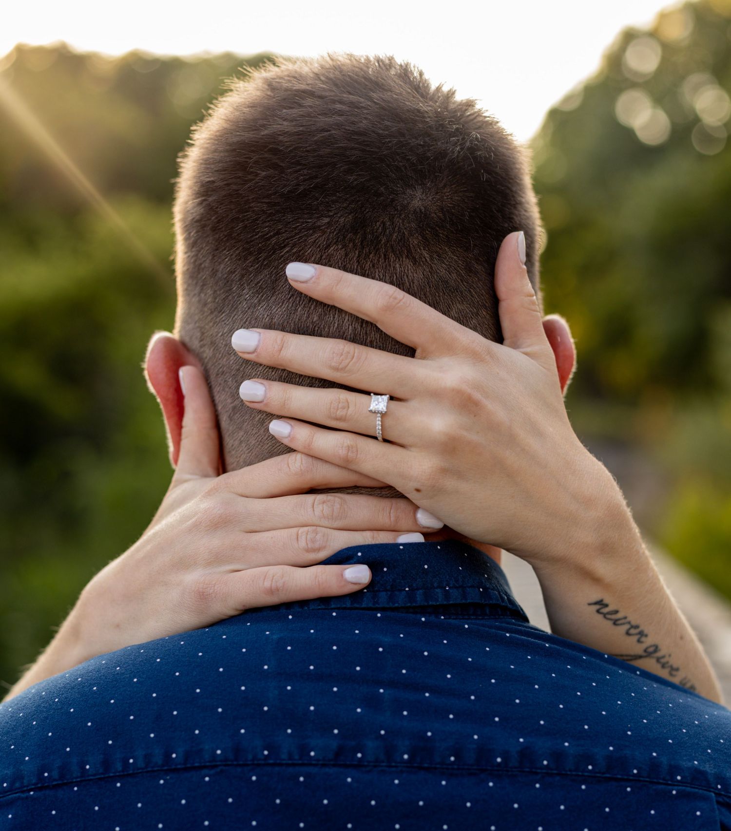 Close-up of engaged woman\u2019s hand with diamond ring resting on her partner\u2019s neck, tattoo visible.