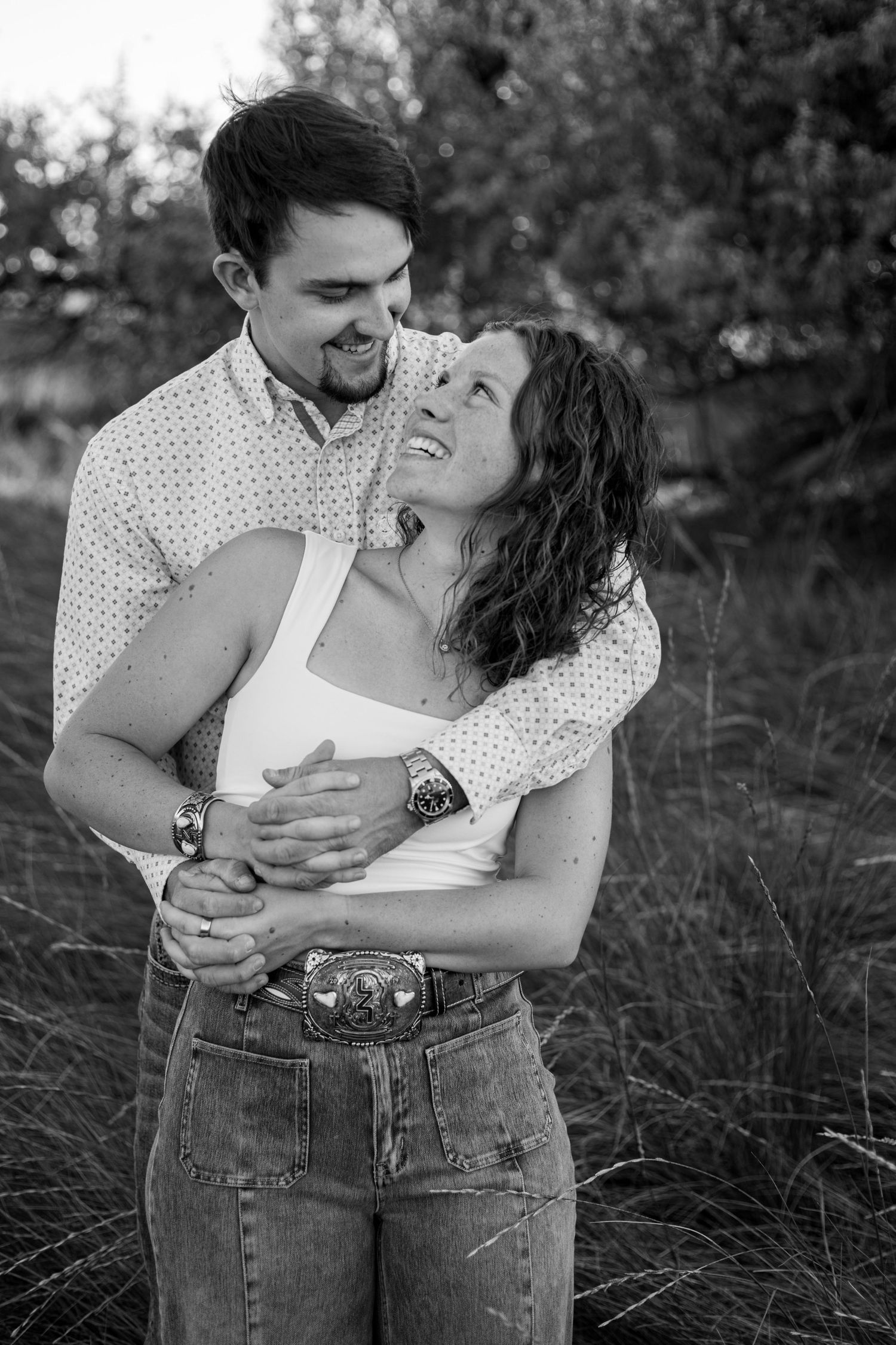 Black and white photo of a couple embracing outdoors, man hugging woman from behind as she smiles up at him.