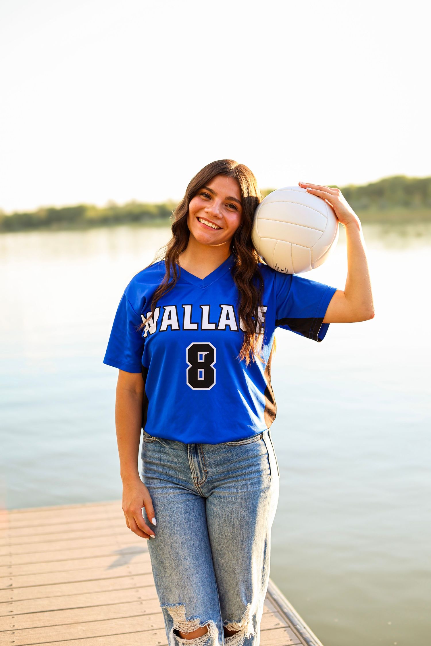 Smiling teen athlete in blue Wallace volleyball jersey holding ball on wooden dock by lake at sunset.