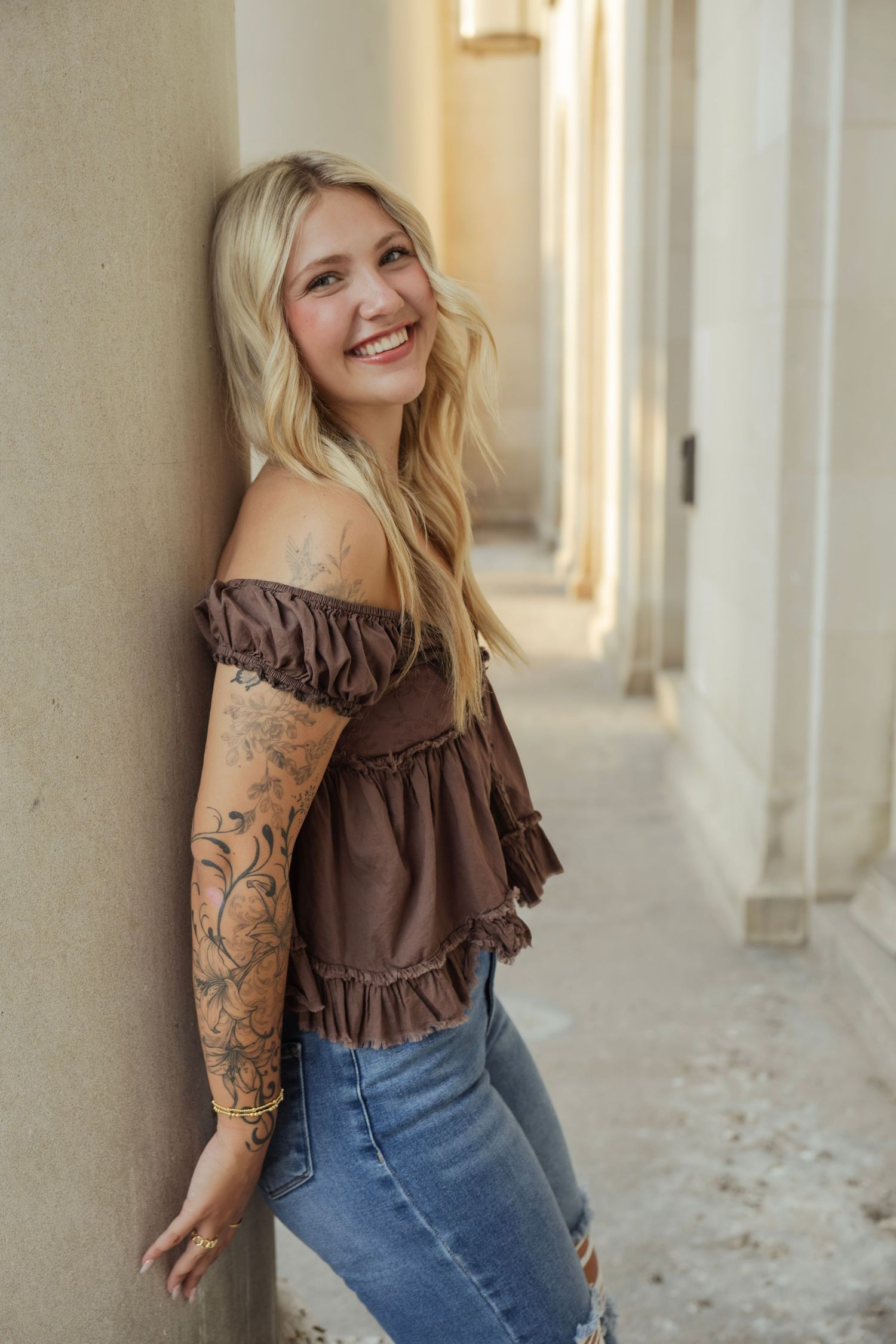 Smiling blonde woman with tattoos wearing brown off-shoulder top and ripped jeans leaning against a wall.