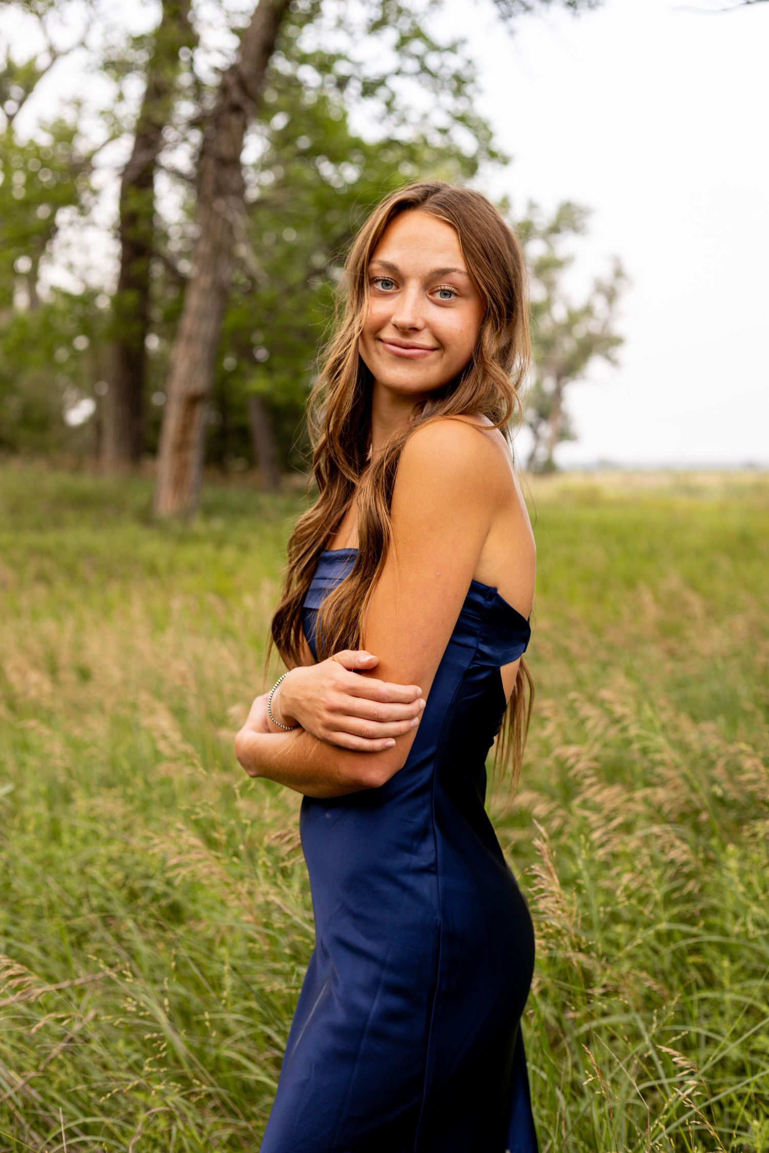 Smiling teenage girl in strapless navy dress poses in tall grass field surrounded by trees.
