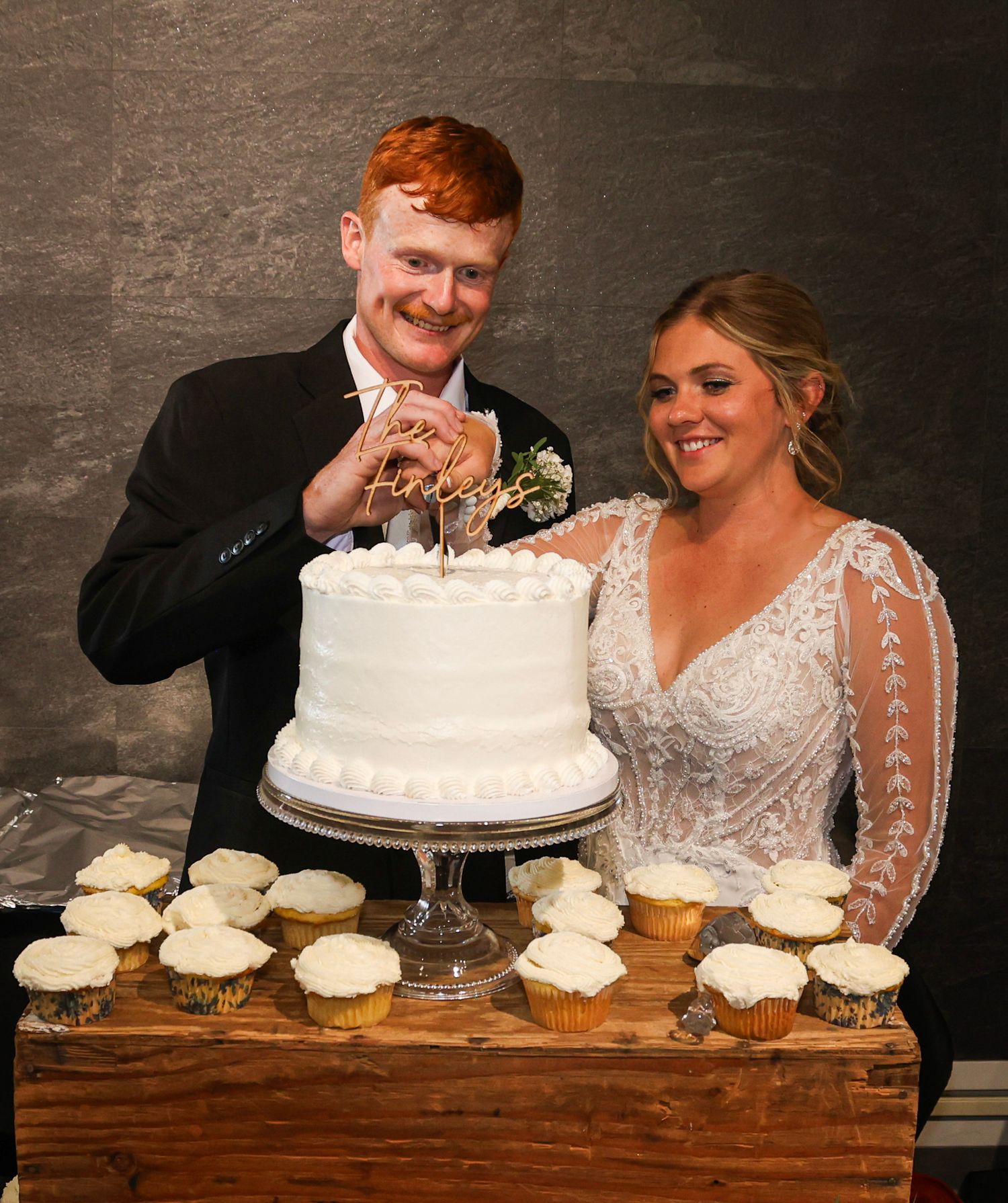 Smiling bride and groom cut white two-tier wedding cake surrounded by cupcakes at their reception.