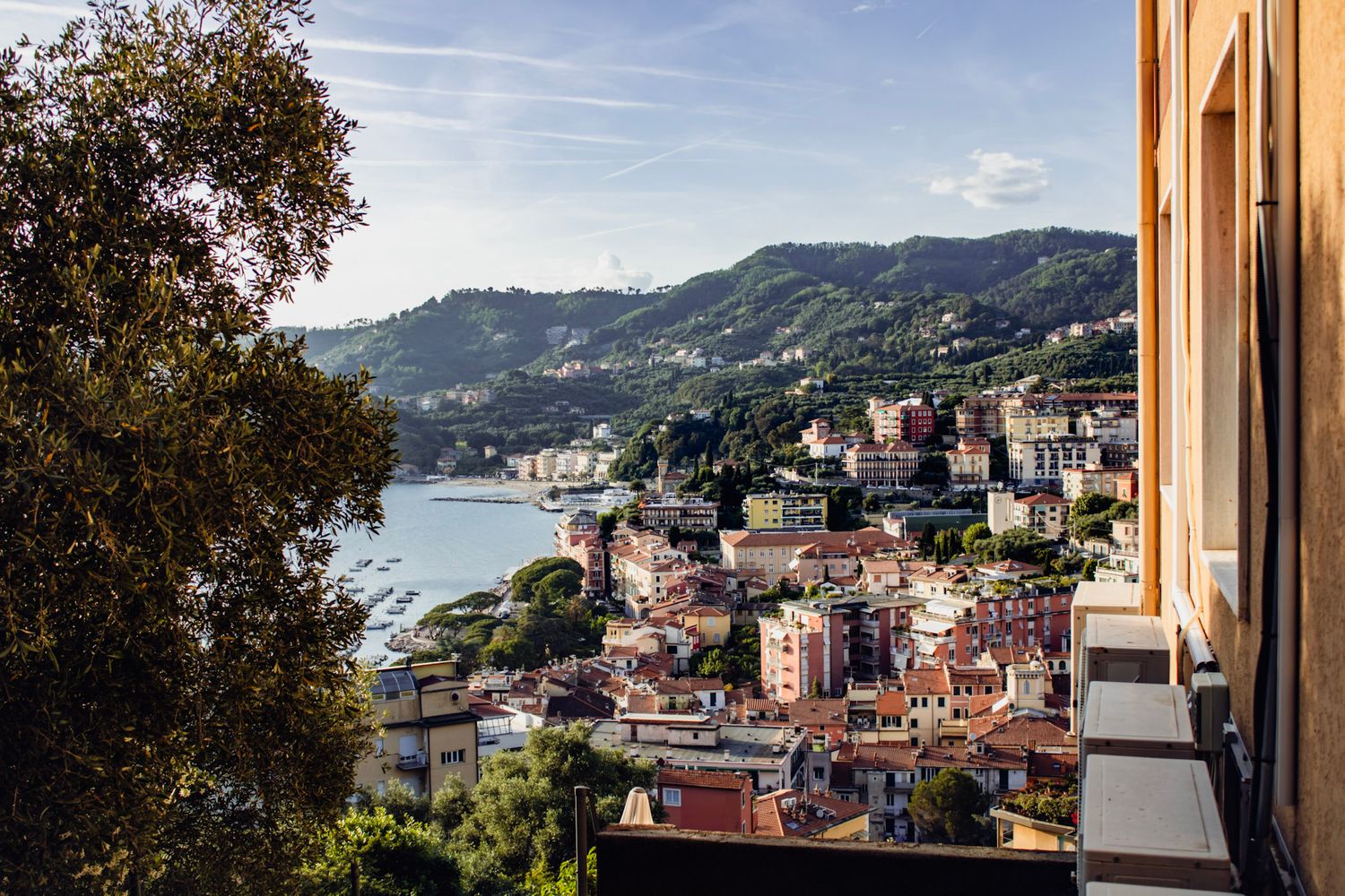 Panoramic view of a colorful Italian coastal town with terracotta rooftops, lush green hills, and a shimmering bay in the background.