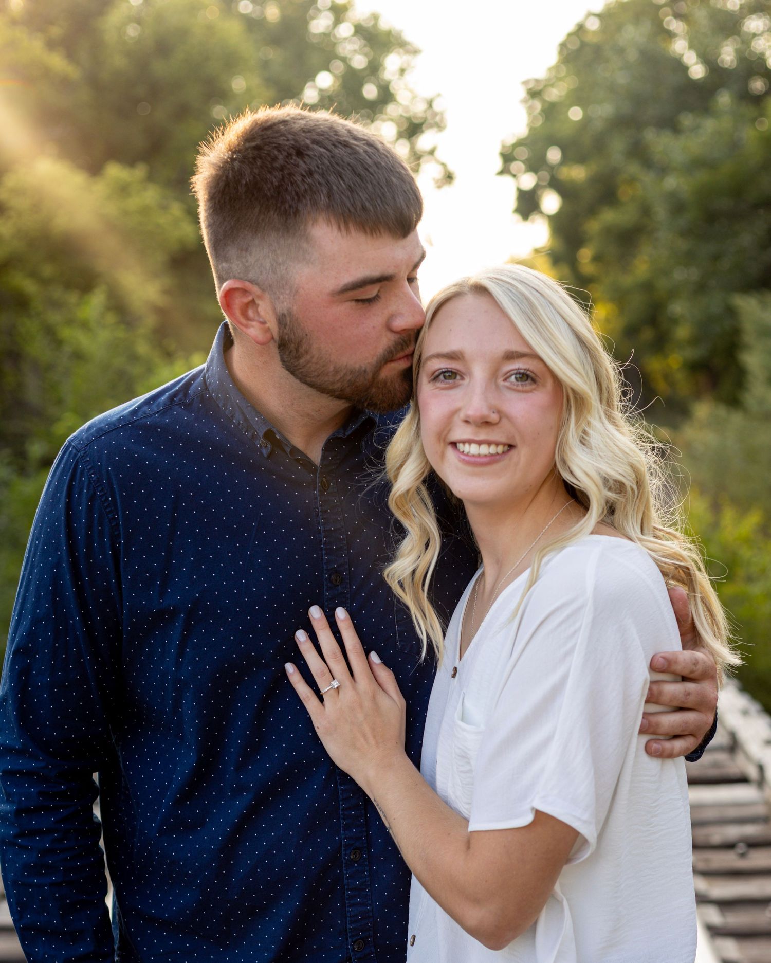 Smiling engaged couple in golden sunset light, woman displaying diamond engagement ring on her hand.