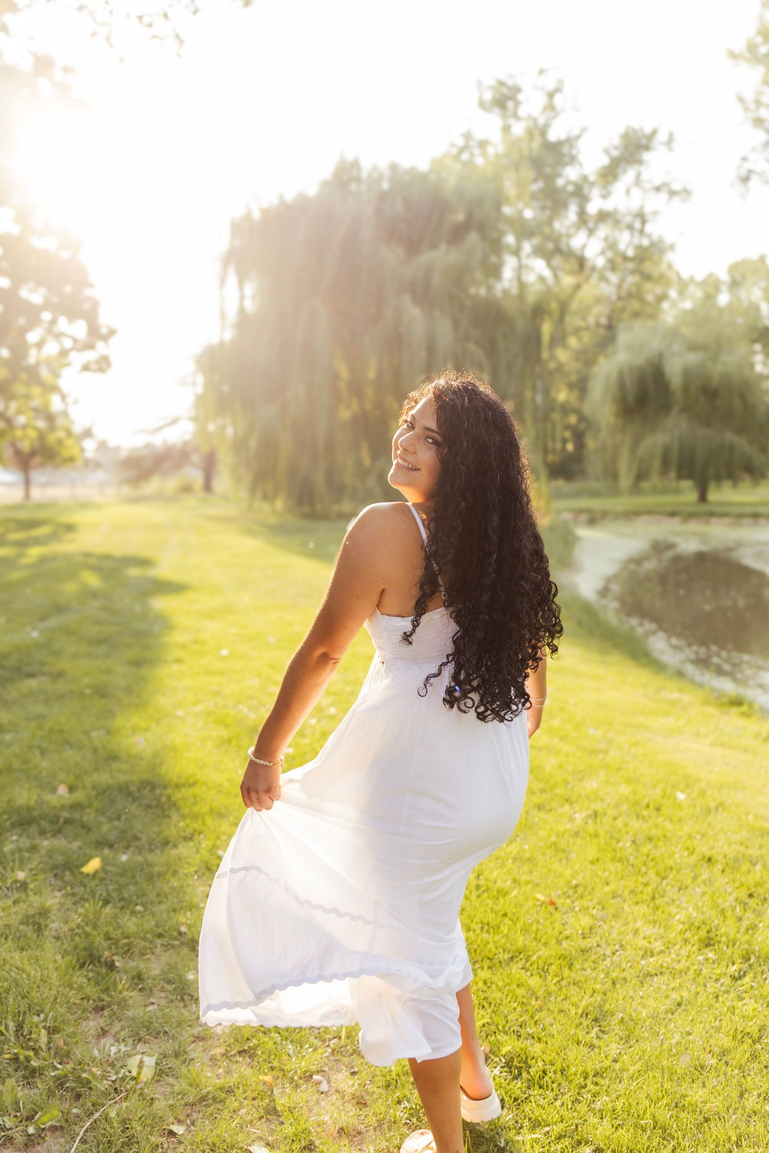 Young woman in white sundress twirling in sunlit park with lush green grass and willow trees.