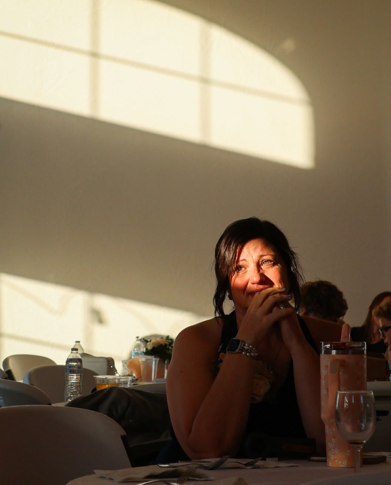 Woman smiling warmly at a banquet table with dramatic light and shadow patterns on the wall behind her.
