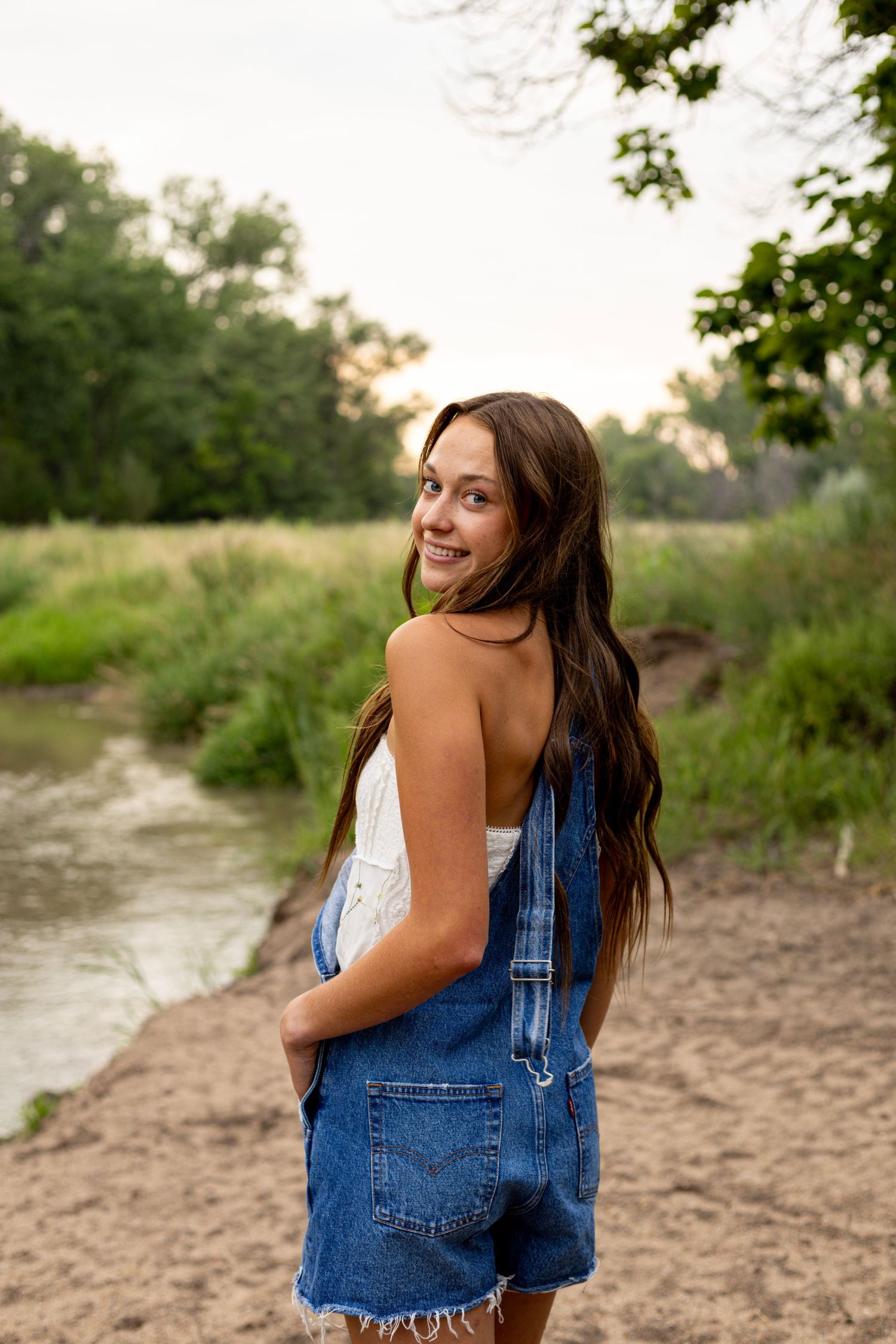 Young woman in denim overalls smiling over her shoulder near a peaceful river surrounded by lush greenery.