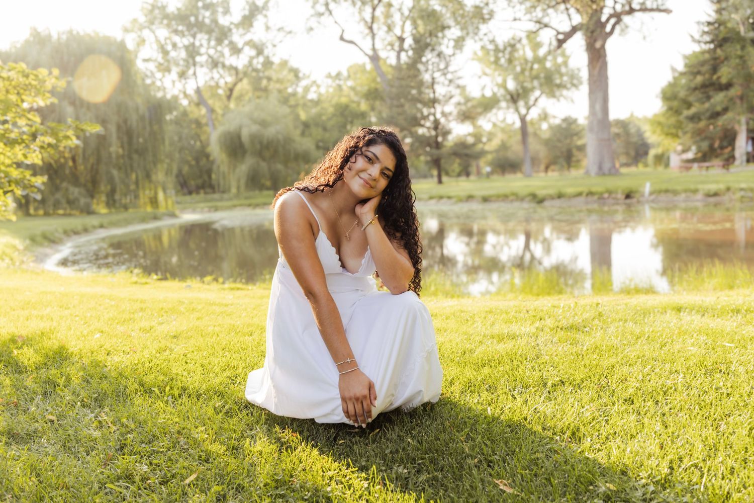 Young woman in white dress sitting on green grass by a pond in a sunlit park.