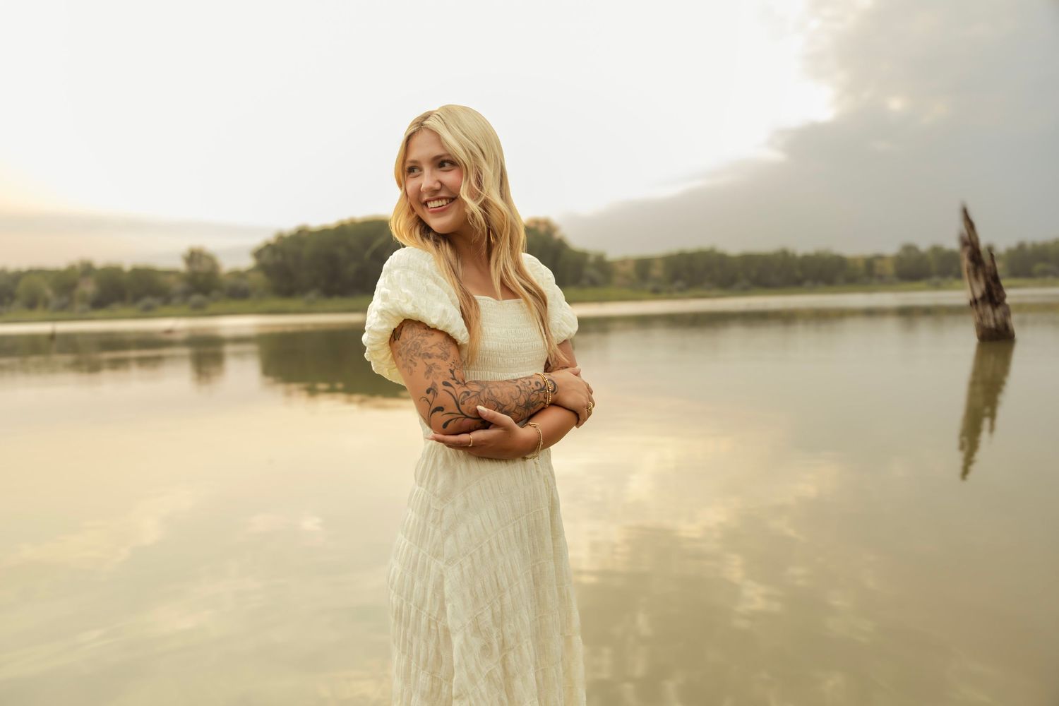 A blonde woman in a white lace dress and fur shawl stands by a serene lake at golden hour.