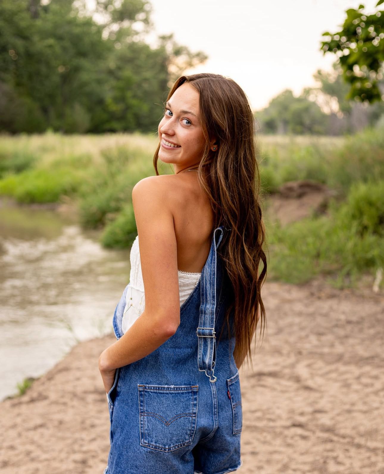 Young woman in Levis denim overalls smiling over shoulder near creek during outdoor senior portrait session.