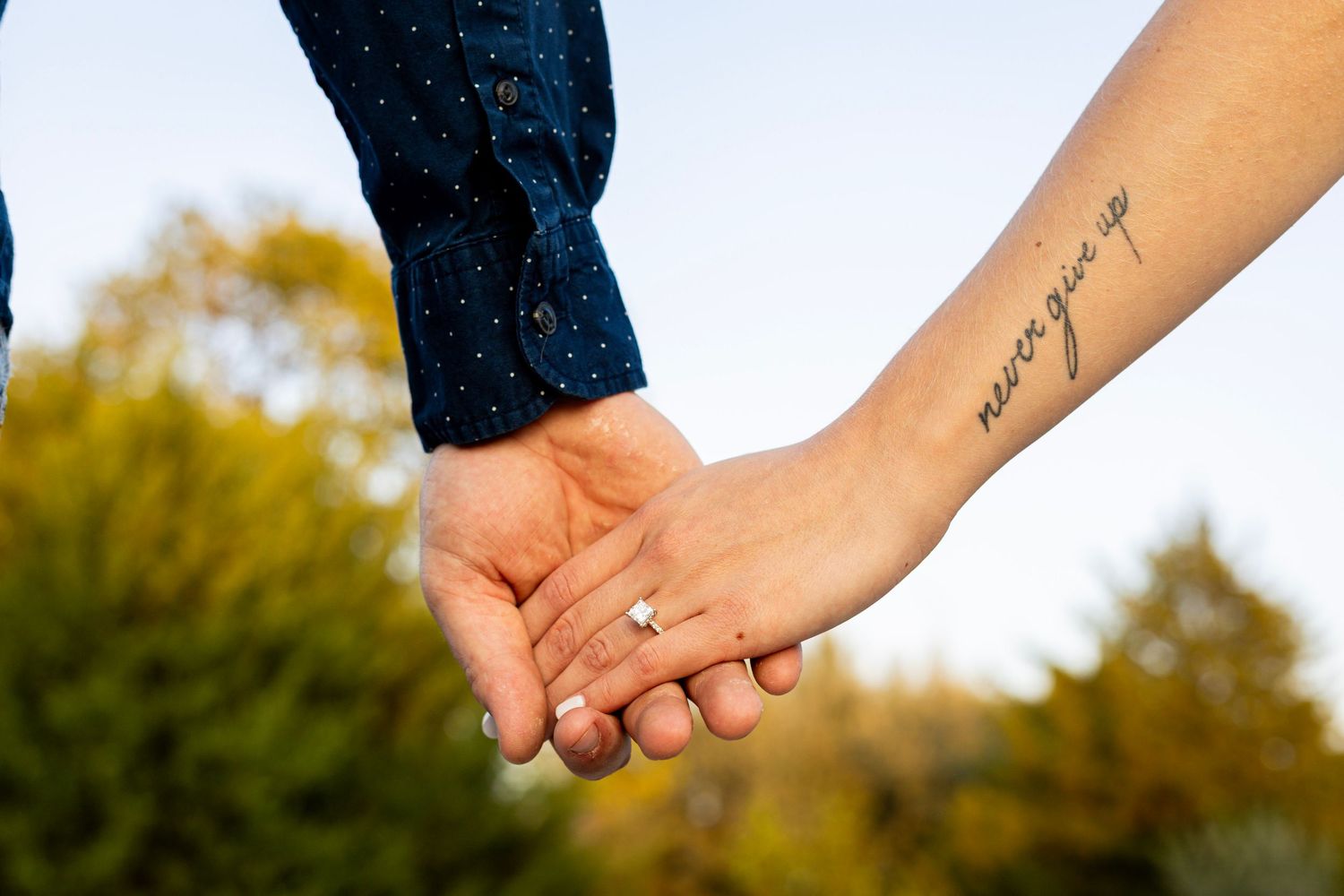 Couple holding hands showing engagement ring and never give up wrist tattoo outdoors.
