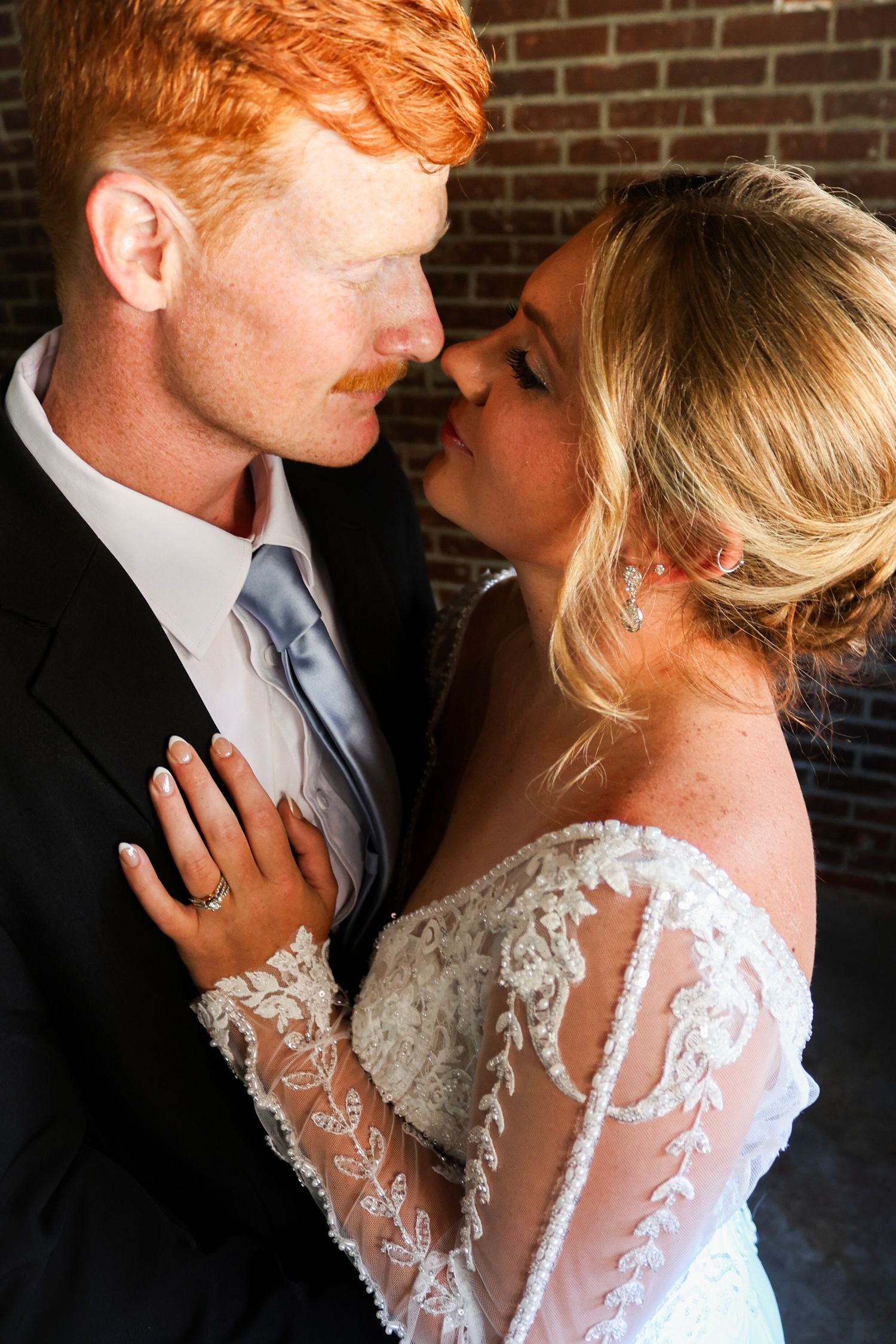 Bride and groom share an intimate nose-to-nose moment against a rustic brick wall backdrop.