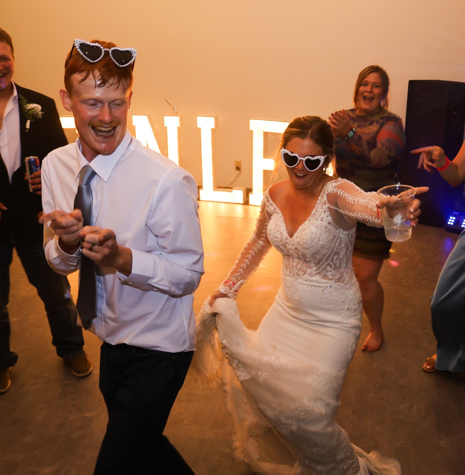 Bride and groom dance joyfully wearing heart-shaped sunglasses with guests at wedding reception.